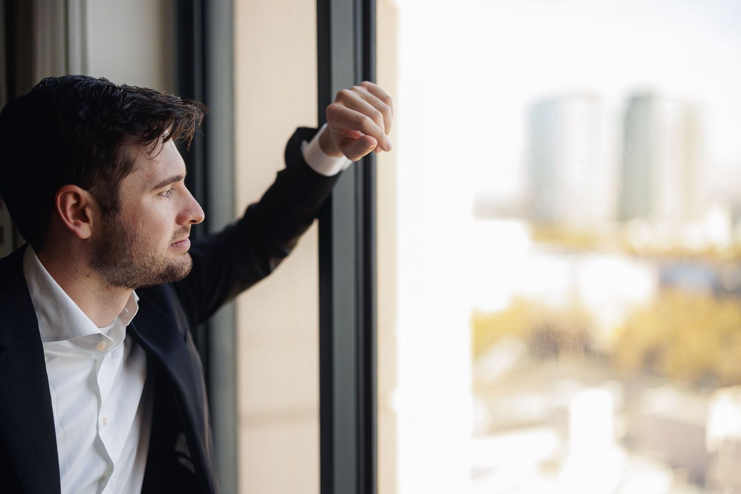 Businessman in dark suit gazes thoughtfully out office window at city skyline on bright day.