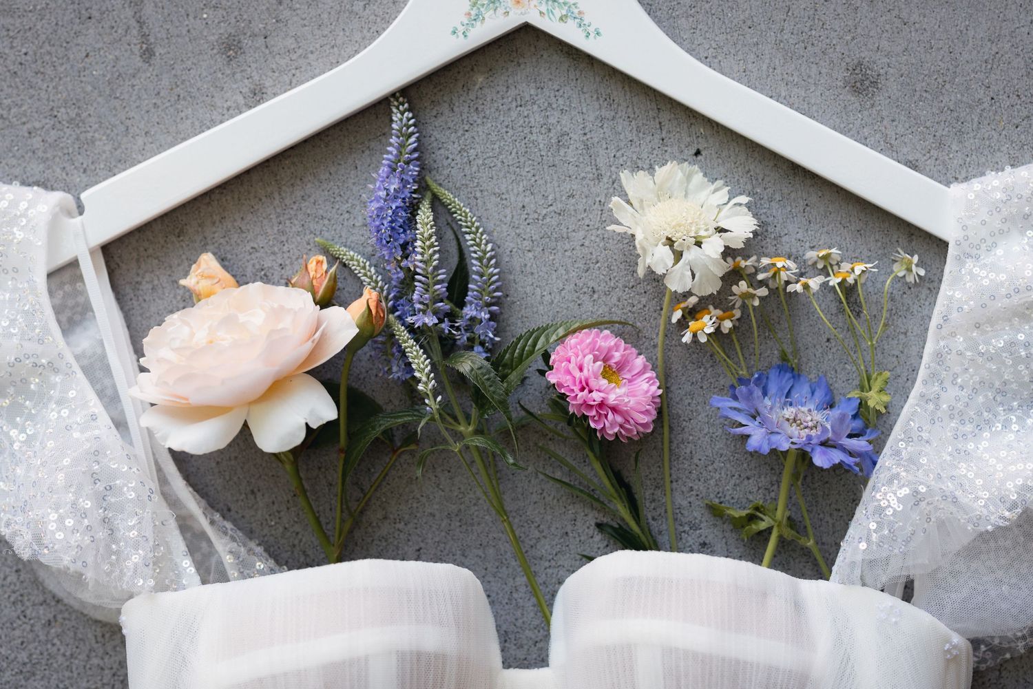 A delicate arrangement of peach rose, pink carnation, blue cornflower, and white flowers against a grey background in a hexagonal frame.