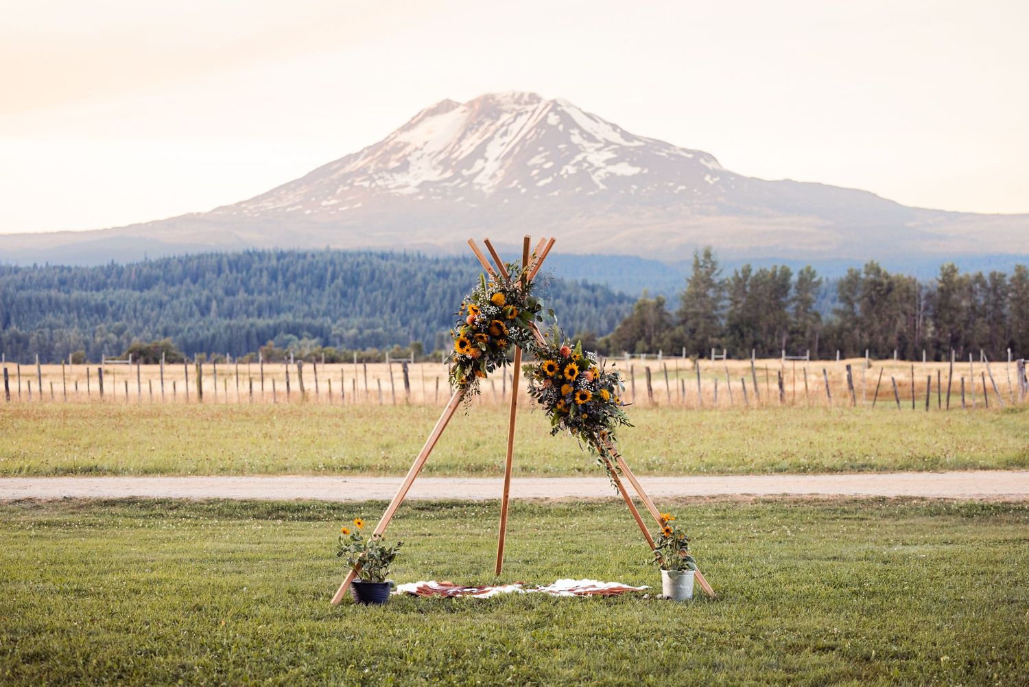 A wooden triangle arch decorated with greenery stands in a field with Mount Adams visible in the distance.