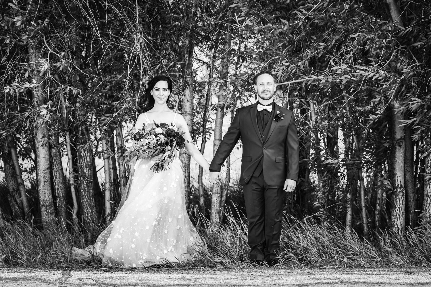 A couple in wedding attire holds hands while standing together in front of a wooded forest backdrop in black and white.
