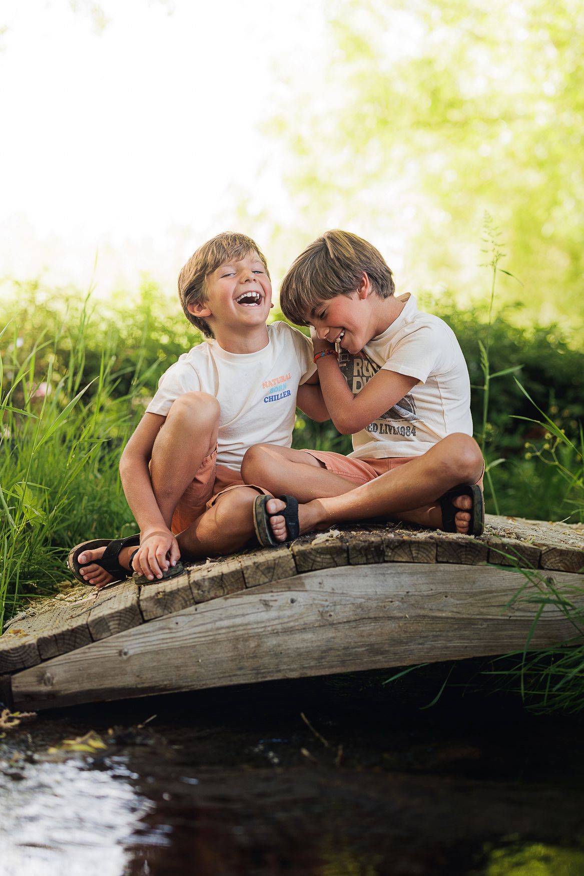 Two children laughing while sitting on wooden bridge over stream.