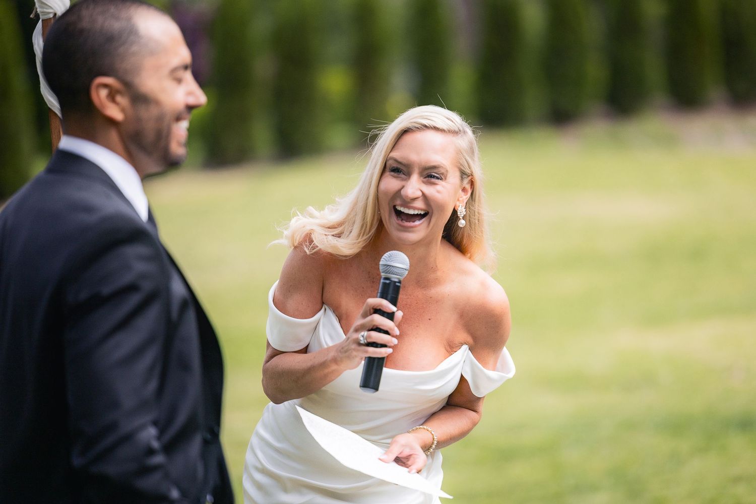 A bride laughs joyfully while holding a microphone during an outdoor wedding ceremony.