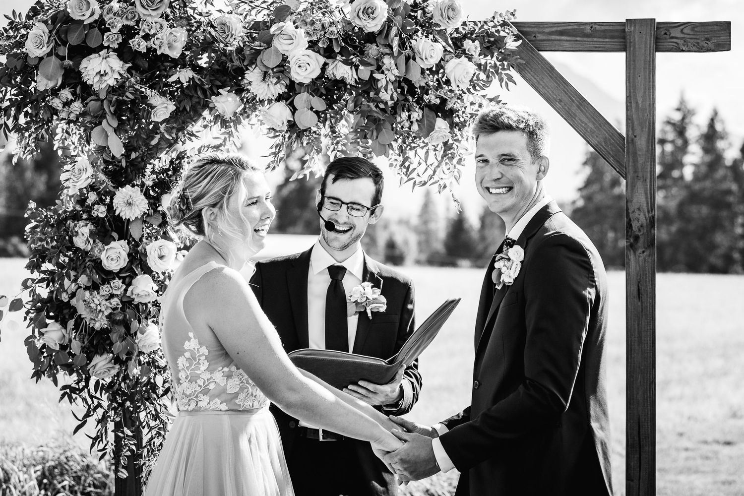 A black and white wedding ceremony under a wooden arch decorated with lush floral arrangements outdoors.