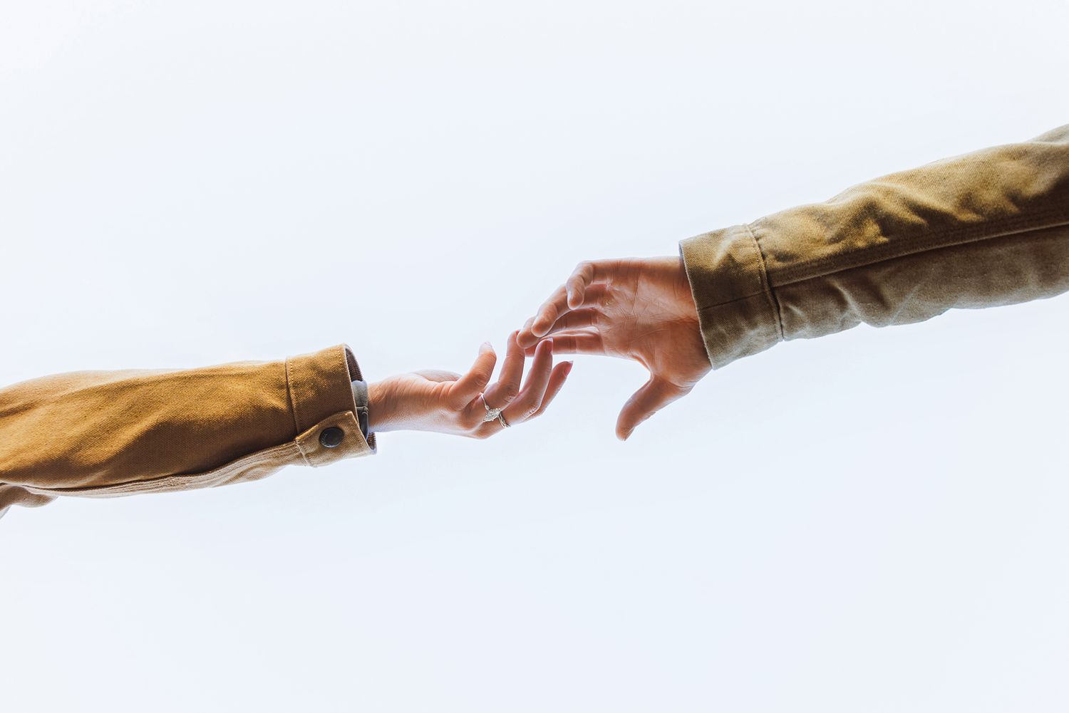 Two outstretched hands in tan jackets reaching toward each other against a bright white sky, nearly touching fingertips.