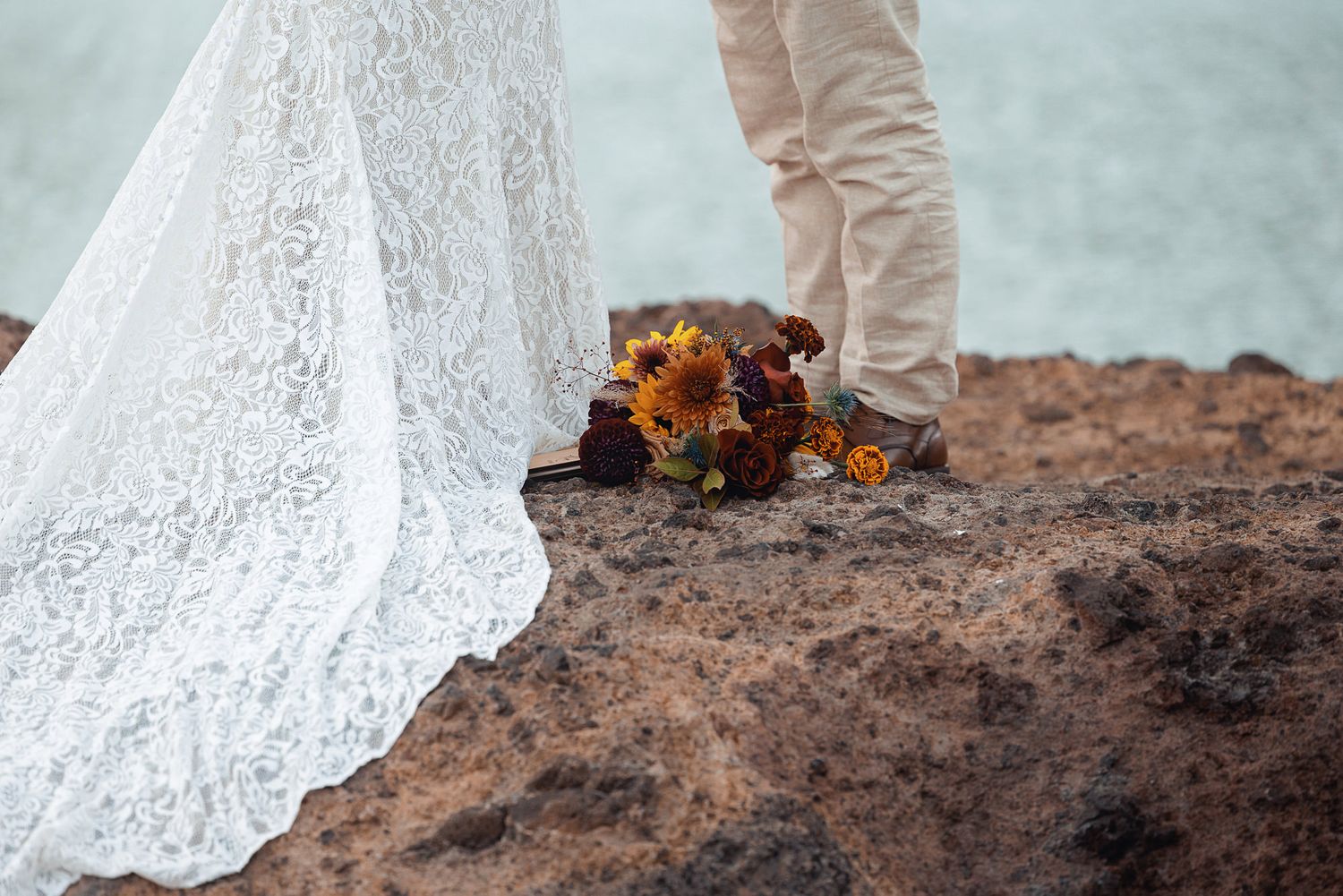 Bride in a flowing white lace wedding gown stands beside a groom in khaki pants on a rocky seaside cliff with an autumn bouquet at their fee
