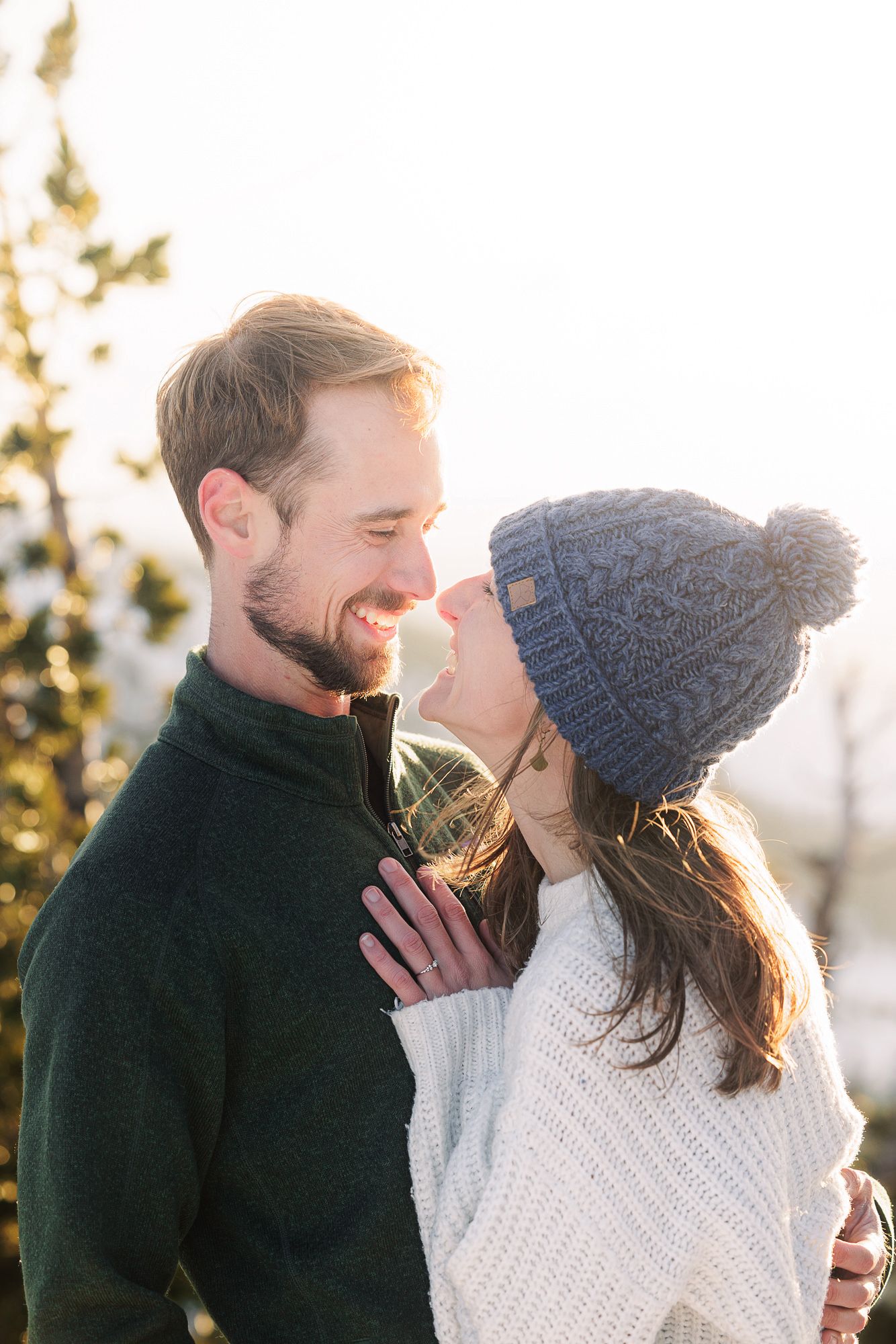 A smiling couple in winter sweaters and a knit hat share a romantic moment outdoors in soft golden sunlight.