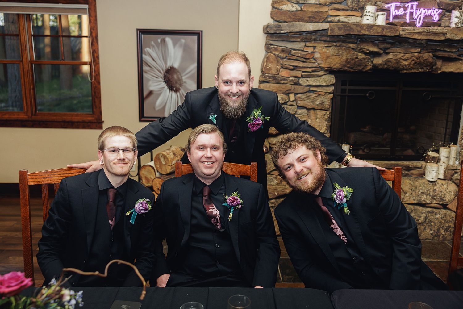 Four men in black suits with floral boutonnieres pose together near a stone fireplace at a wedding reception.