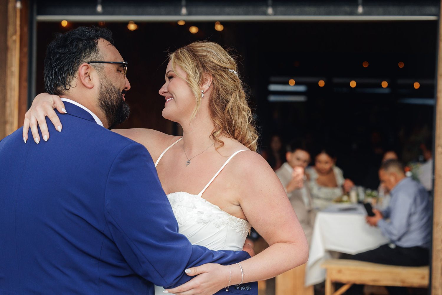 Bride and groom share first dance outdoors at wedding reception, smiling at each other in blue suit and white gown.