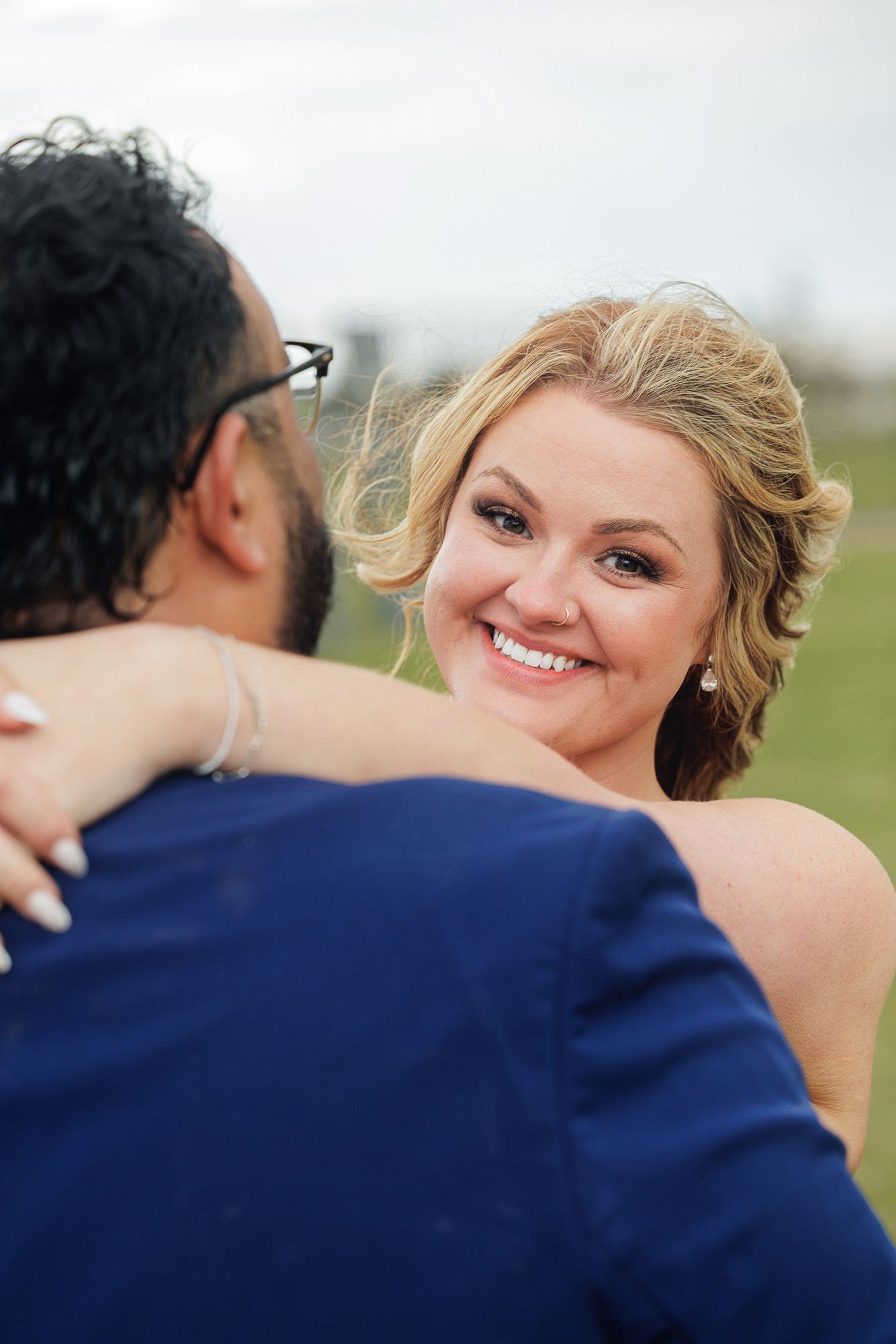 Smiling blonde bride embracing groom in blue suit outdoors on green field during wedding celebration.