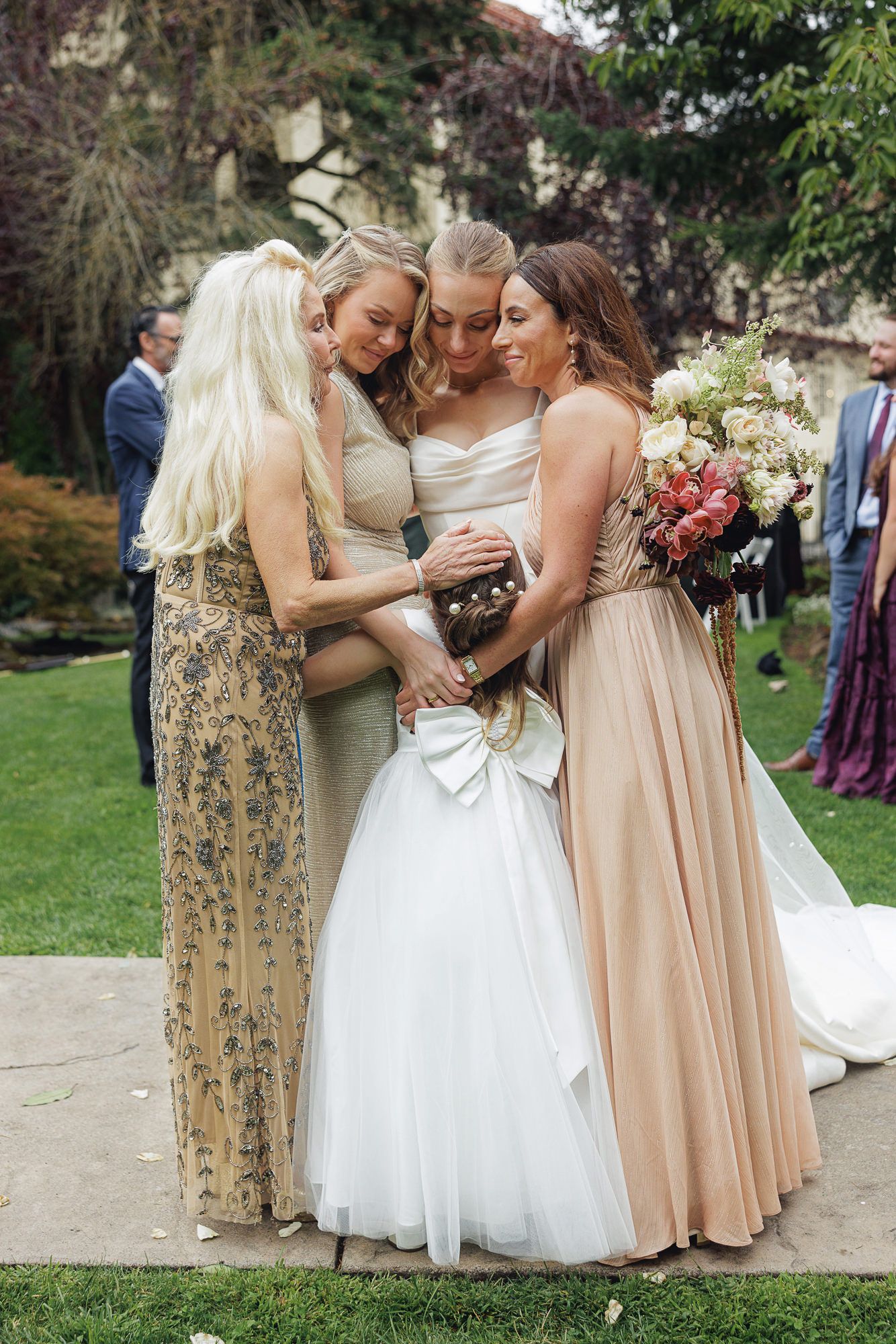 Bride in white gown embraced by two bridesmaids in gold and blush dresses during outdoor wedding ceremony.