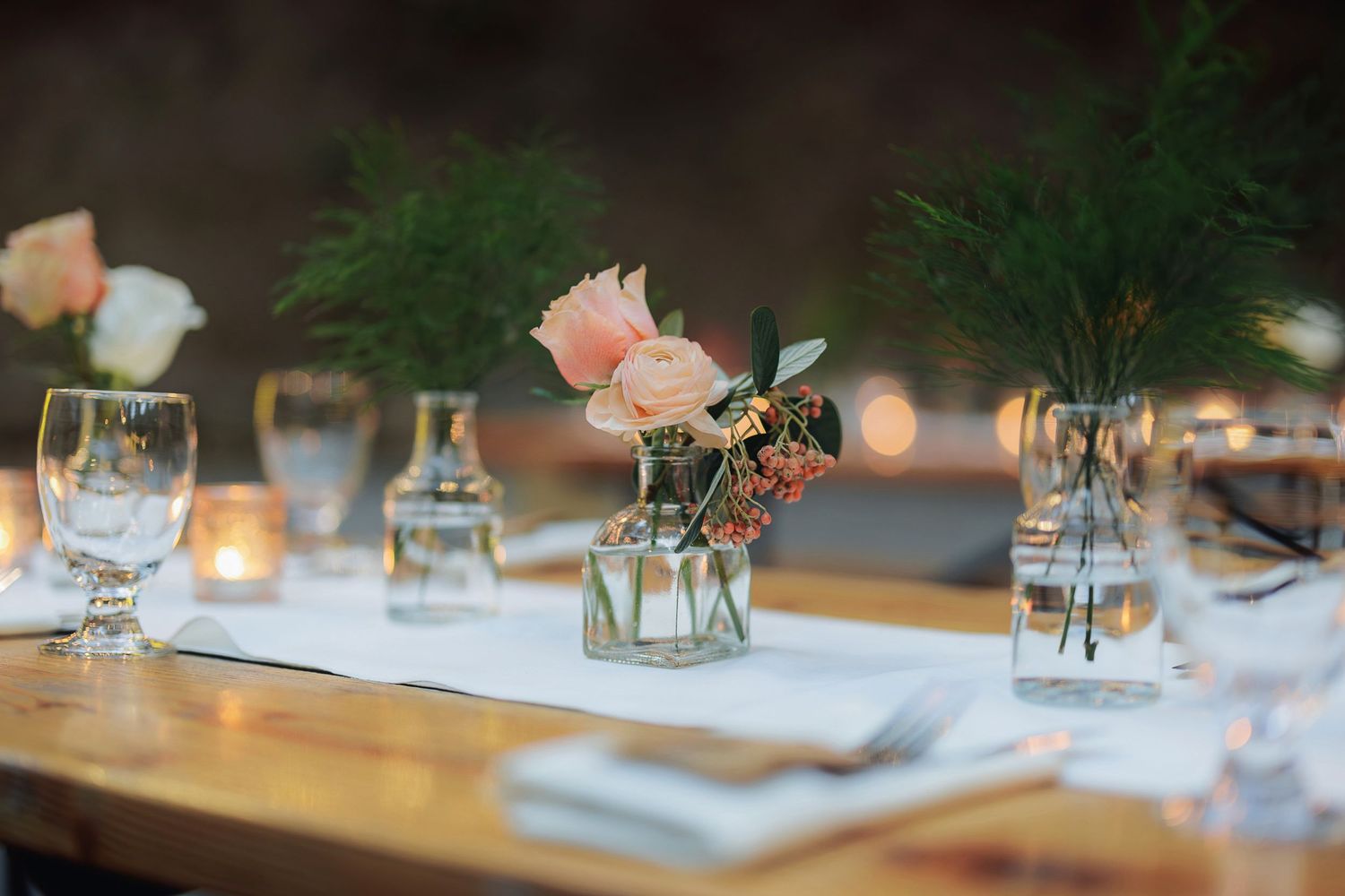Elegant table setting with peach roses in small vases, wine glasses, and candlelight creating a romantic ambiance.
