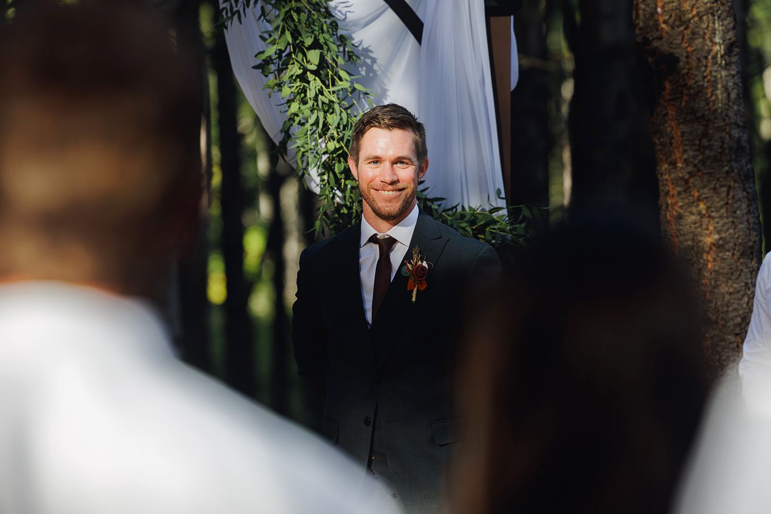 A couple in formal wedding attire embraces in a romantic outdoor setting with trees and natural light.