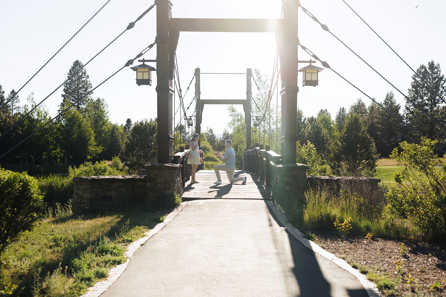 Two people walk across a charming suspension bridge surrounded by lush green trees in a sunny park setting.