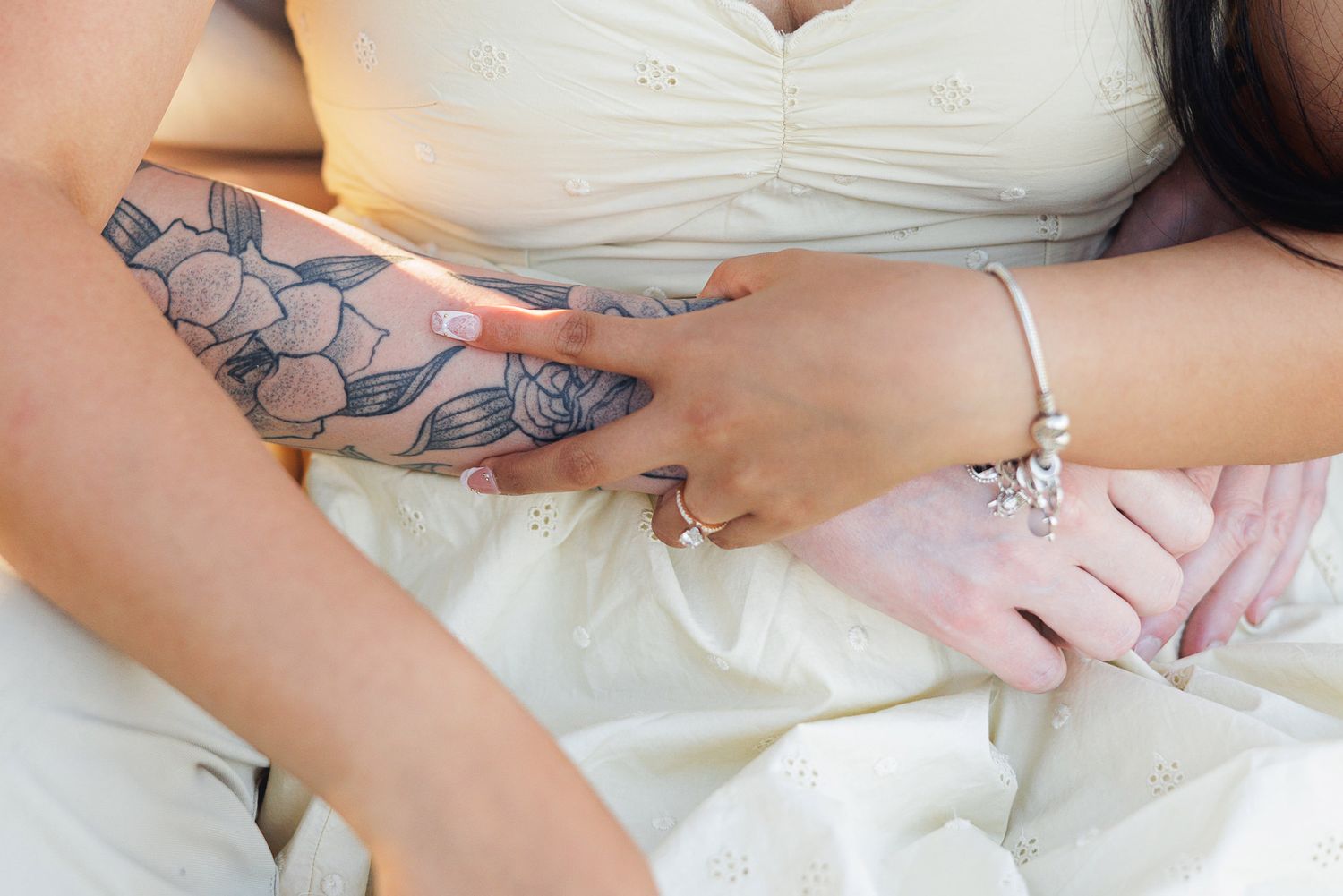 Two women holding hands, one with a floral arm tattoo and bracelet, wearing white dresses in an intimate moment.