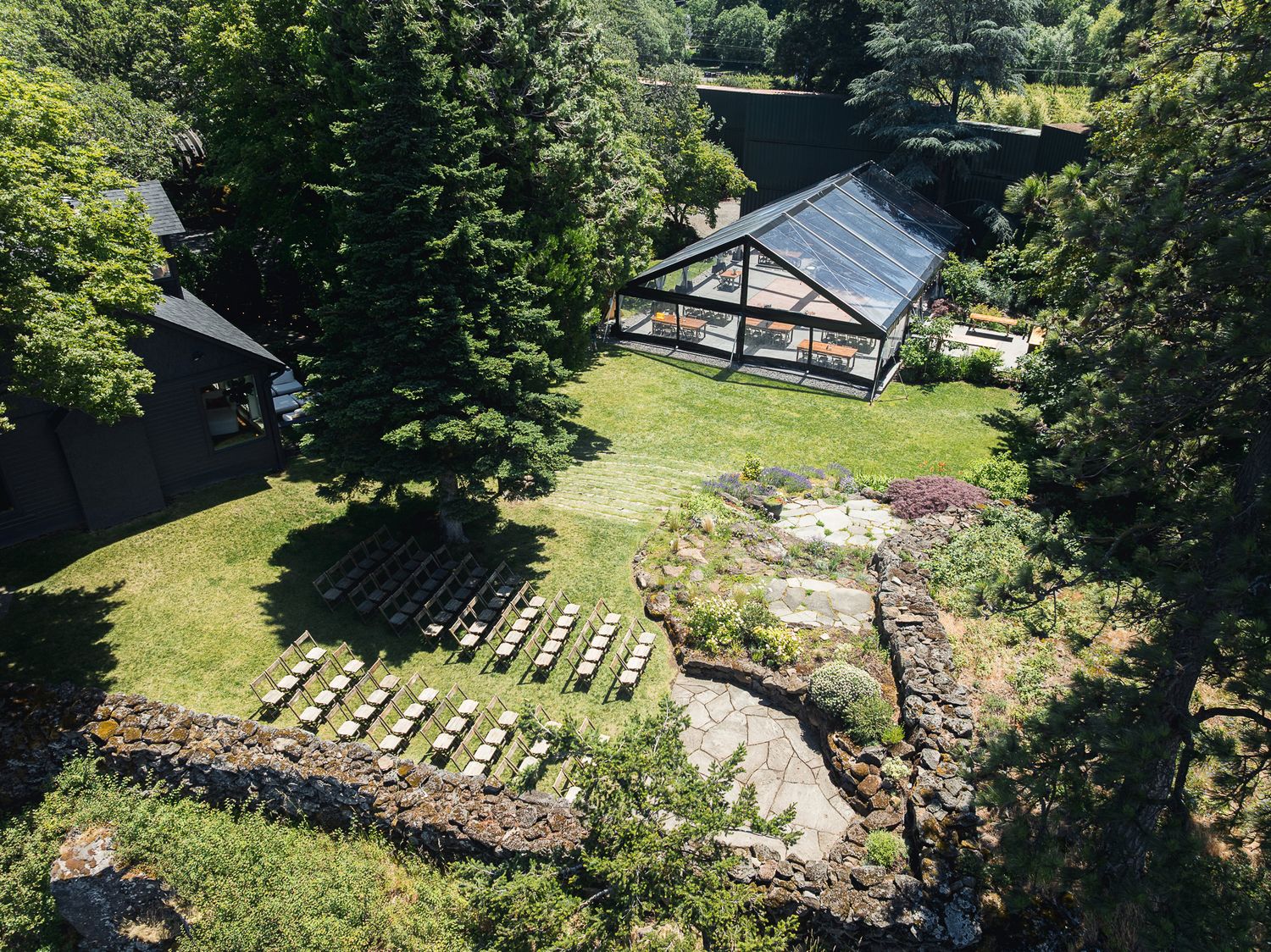 Aerial view of a greenhouse and garden with stone walls, manicured lawn, and rows of plants surrounded by lush trees.