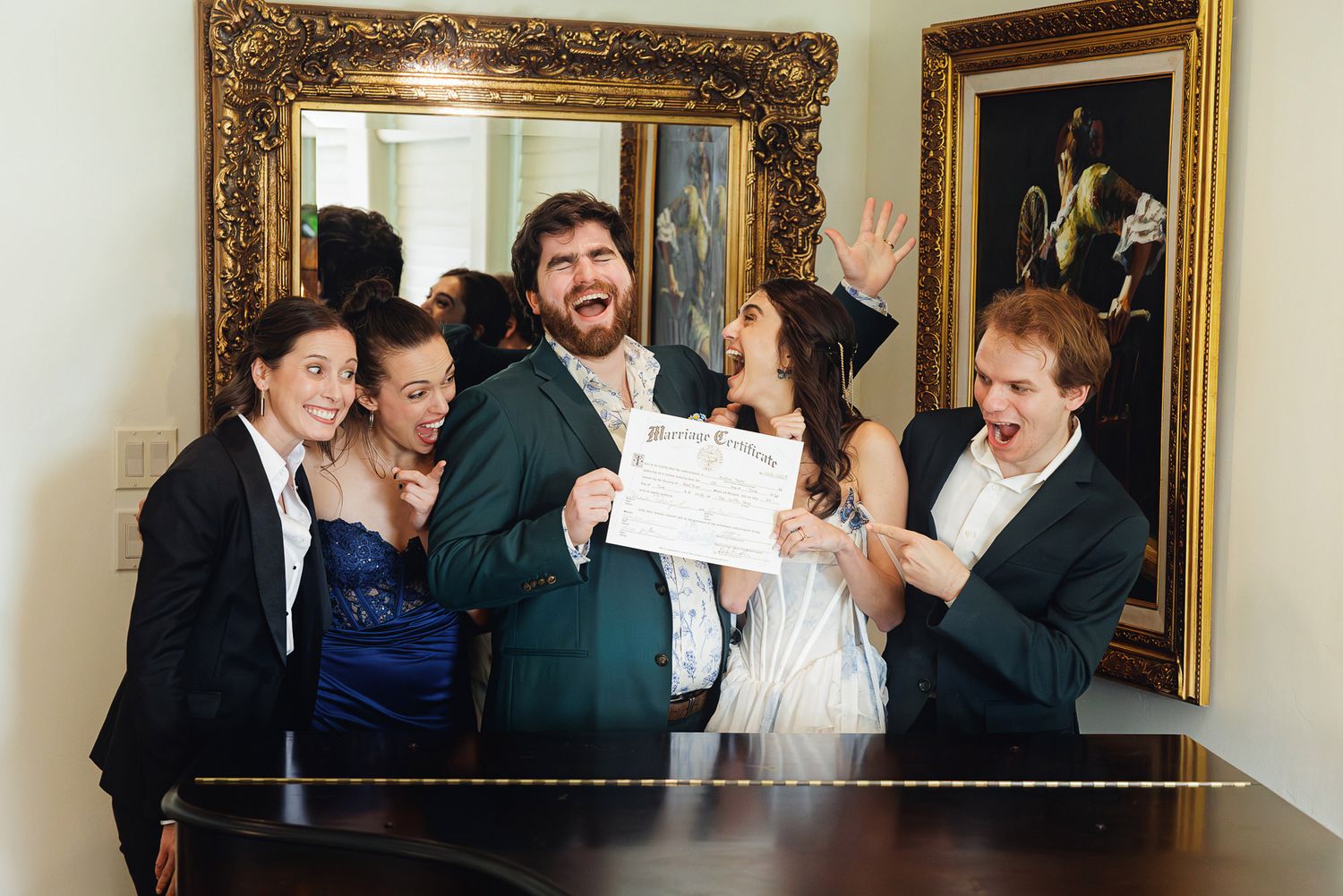 Group of five people celebrating with a certificate, smiling and cheering in front of ornate gold-framed artwork.