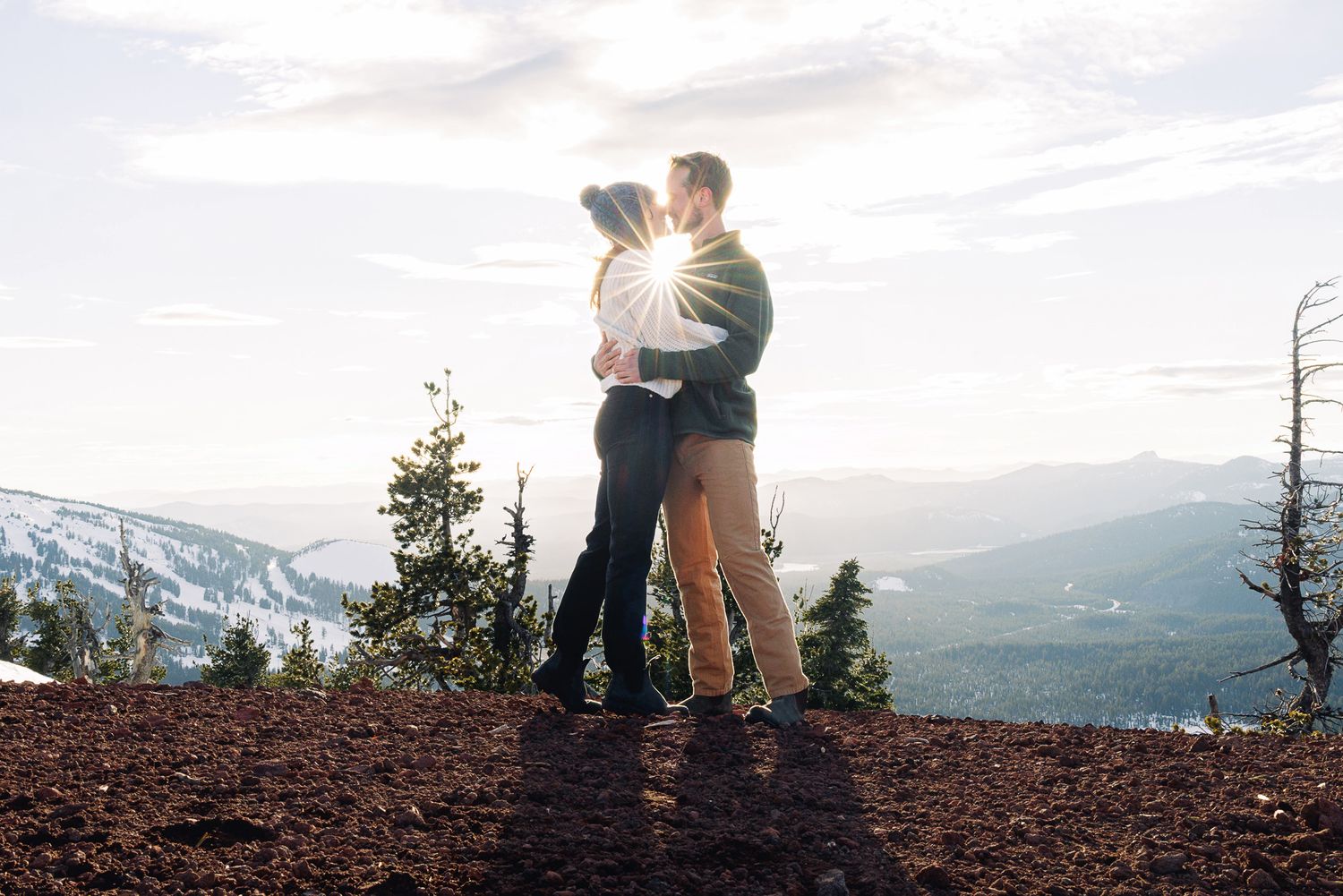Couple kissing on a mountain summit at sunset with panoramic snowy landscape views behind them.