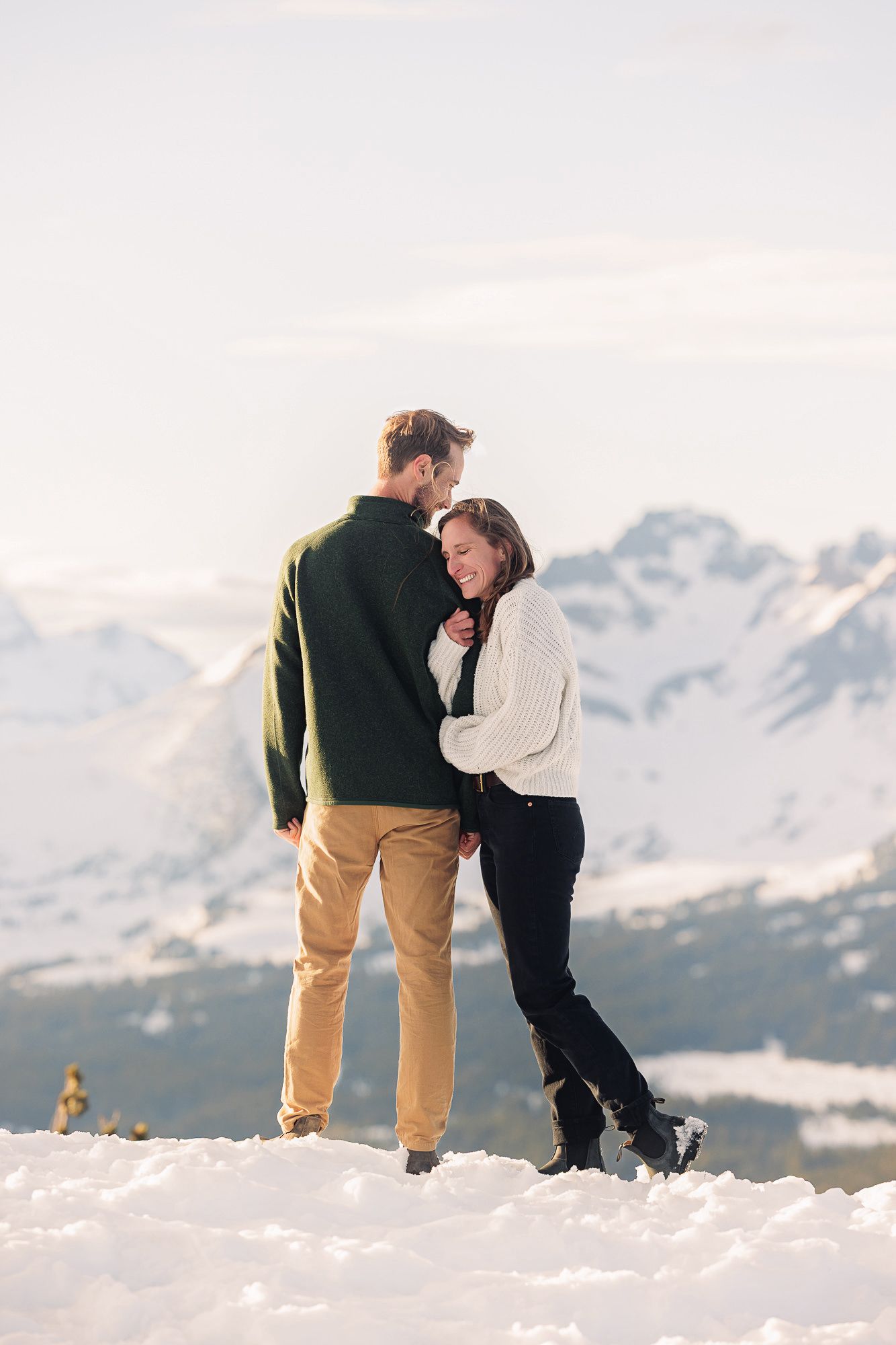 Couple embracing in snowy mountain landscape, wearing cozy sweaters, with scenic snow-capped peaks in background.