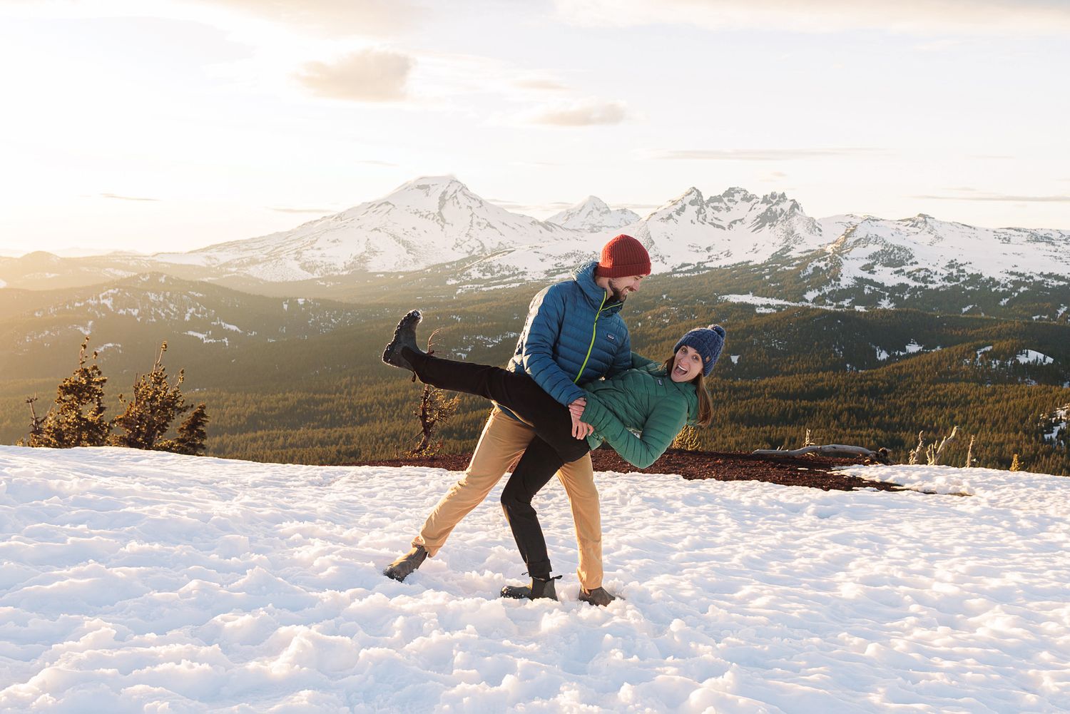 Couple playfully dancing on snowy mountain slope at sunset with snow-capped peaks in background.