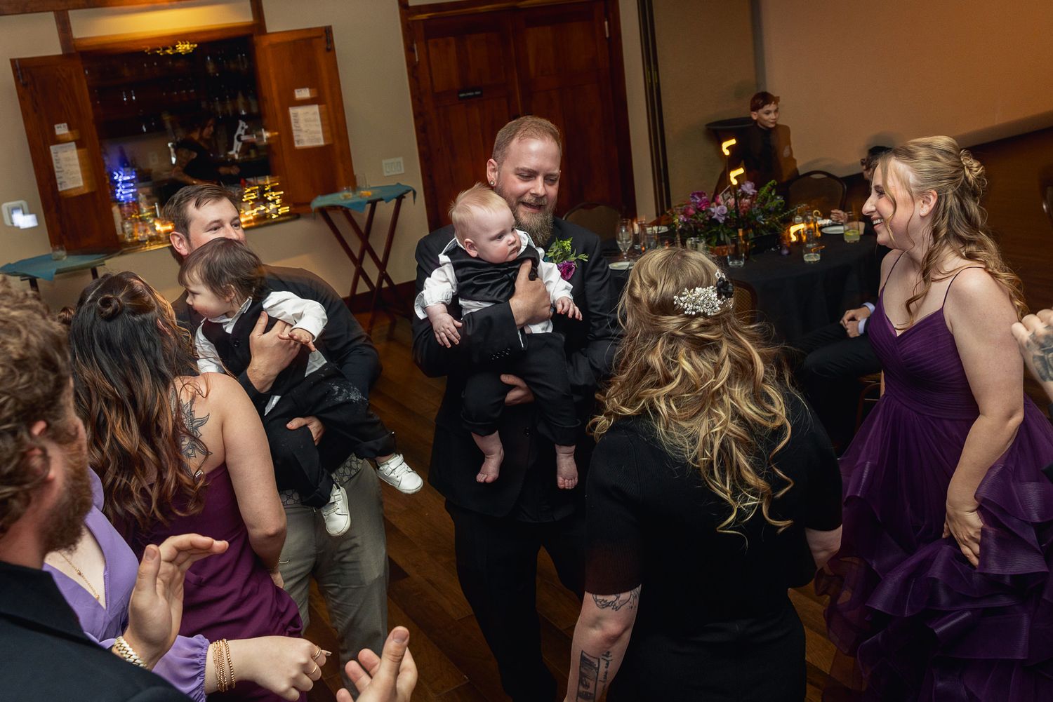 Man in tuxedo holds baby at lively wedding reception dance floor surrounded by guests in purple bridesmaid dresses.