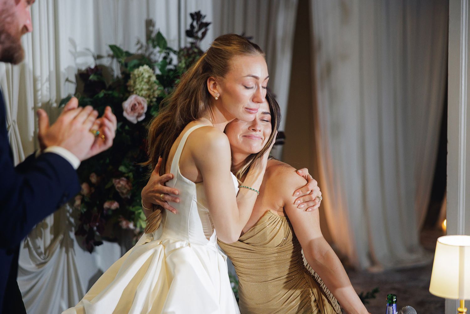 Bride in white gown hugging woman in gold dress at wedding reception with floral arrangements in background.