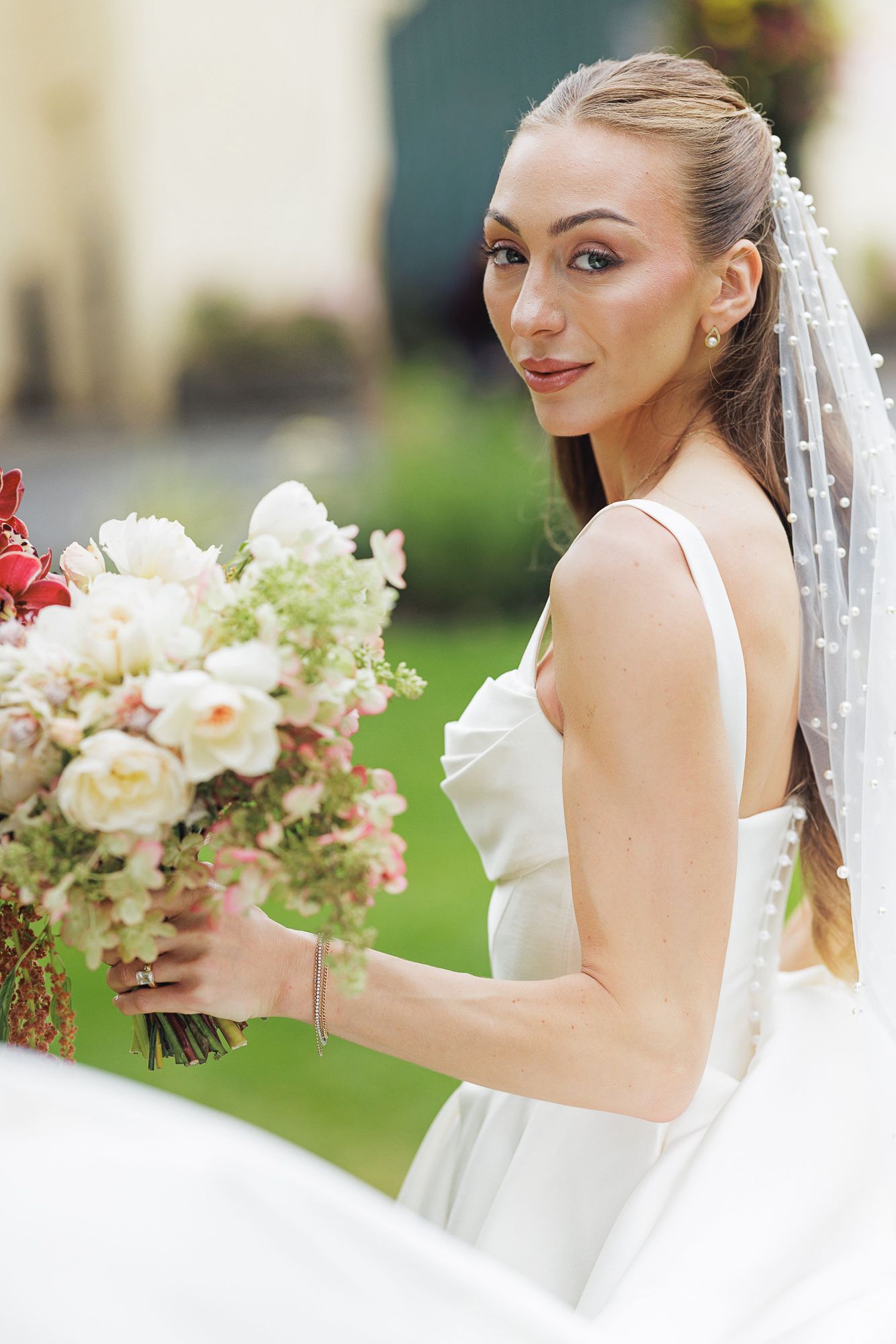 Elegant bride in white dress holding a lush floral bouquet with pink and white flowers, wearing a pearl-adorned veil outdoors.