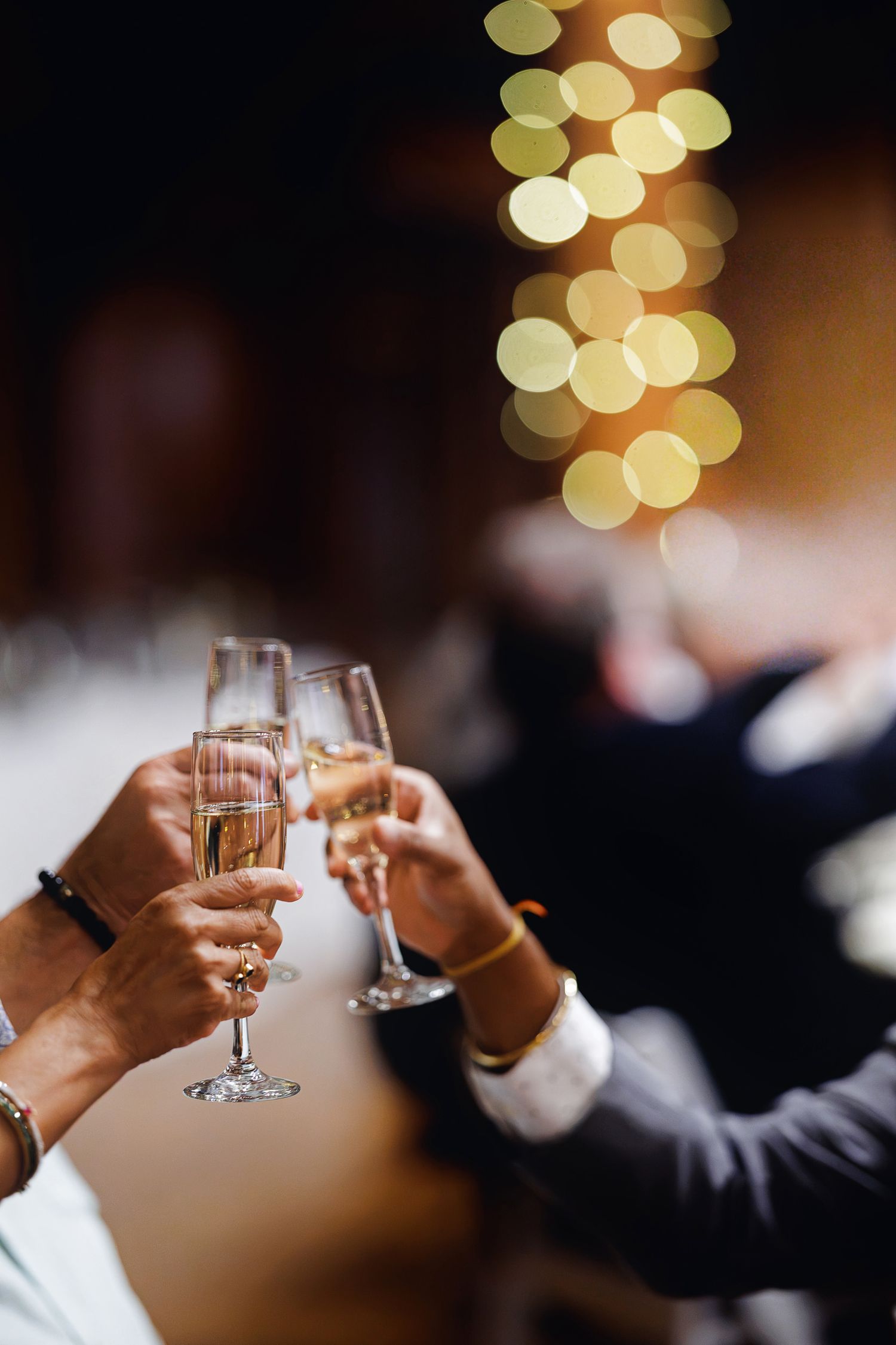 People clinking champagne flutes at a celebration with bokeh lights in the background.
