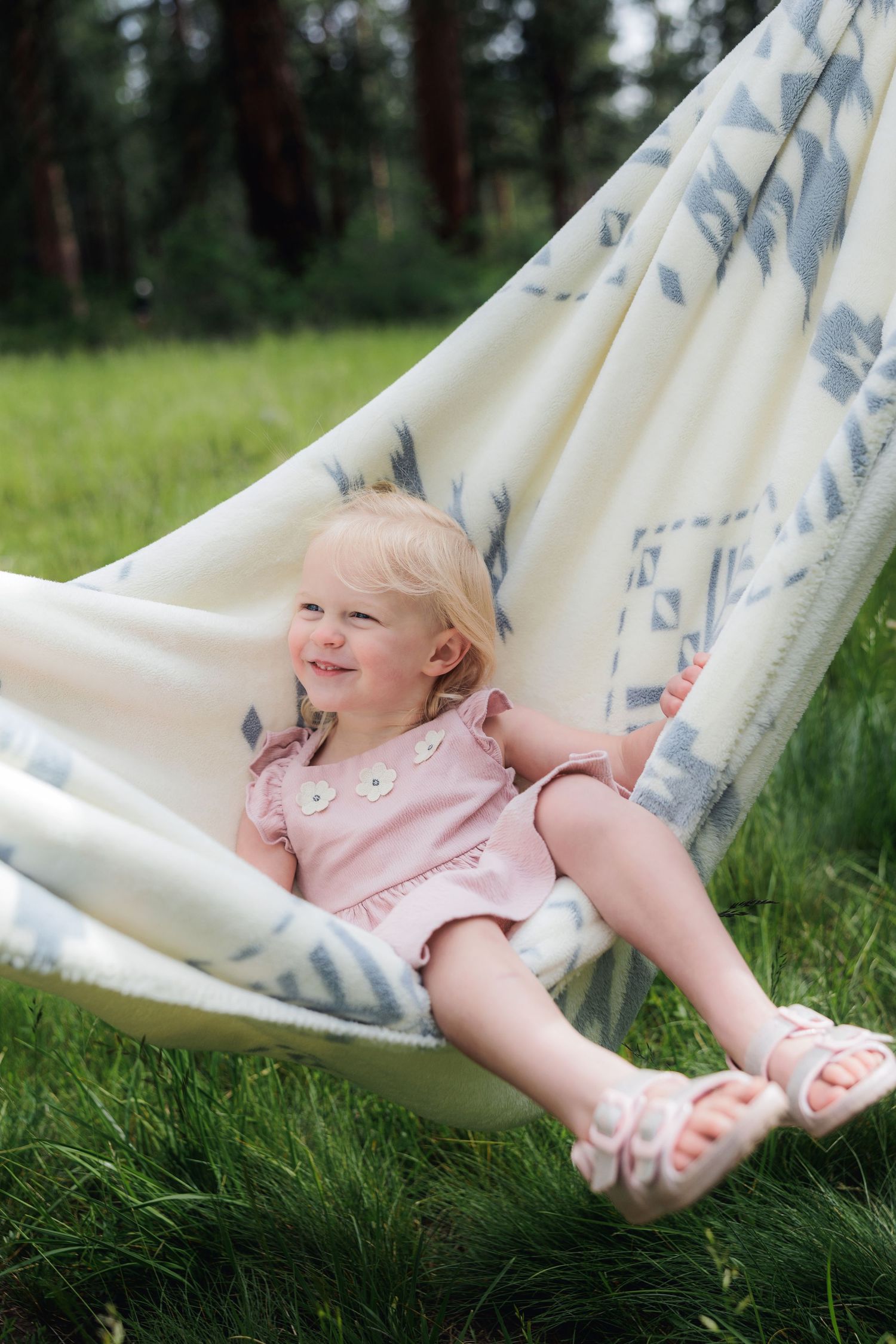 A young child in a pink dress relaxes in a white hammock with blue patterns against a green forest background.