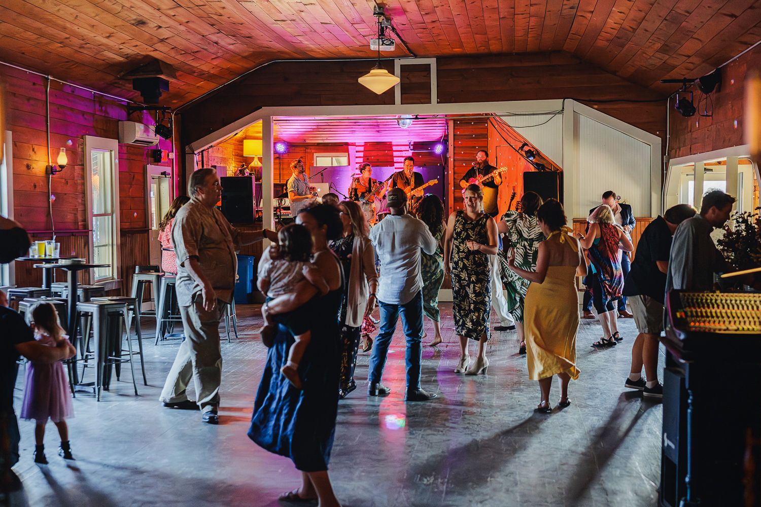 People dance and socialize in a rustic indoor venue illuminated by colorful mood lighting.