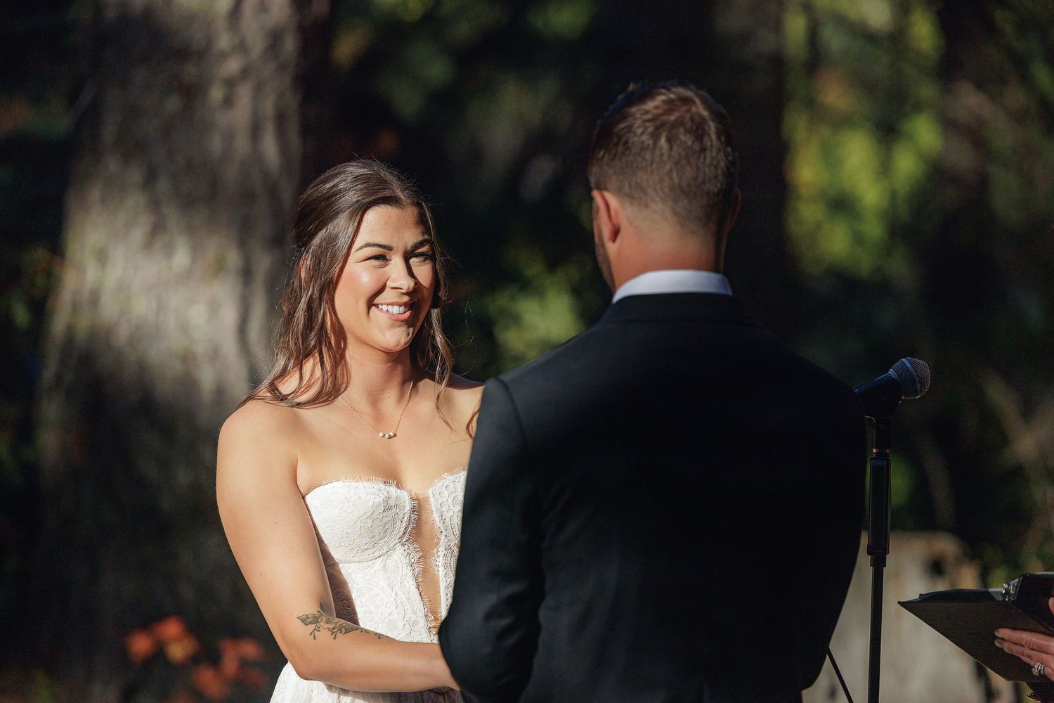 Bride smiling joyfully at groom during outdoor wedding ceremony, wearing strapless white gown among lush green trees.