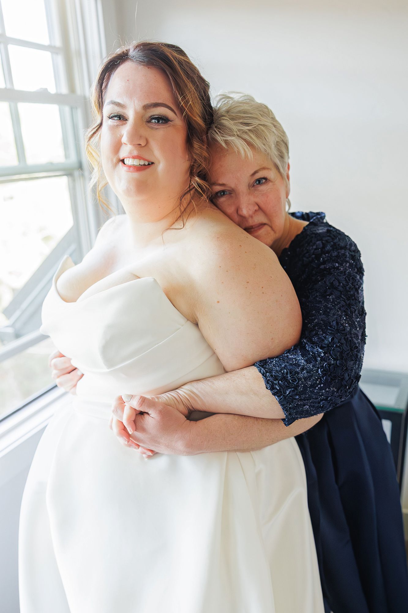 A tender embrace is shared between two people in formal attire against a bright window backdrop.