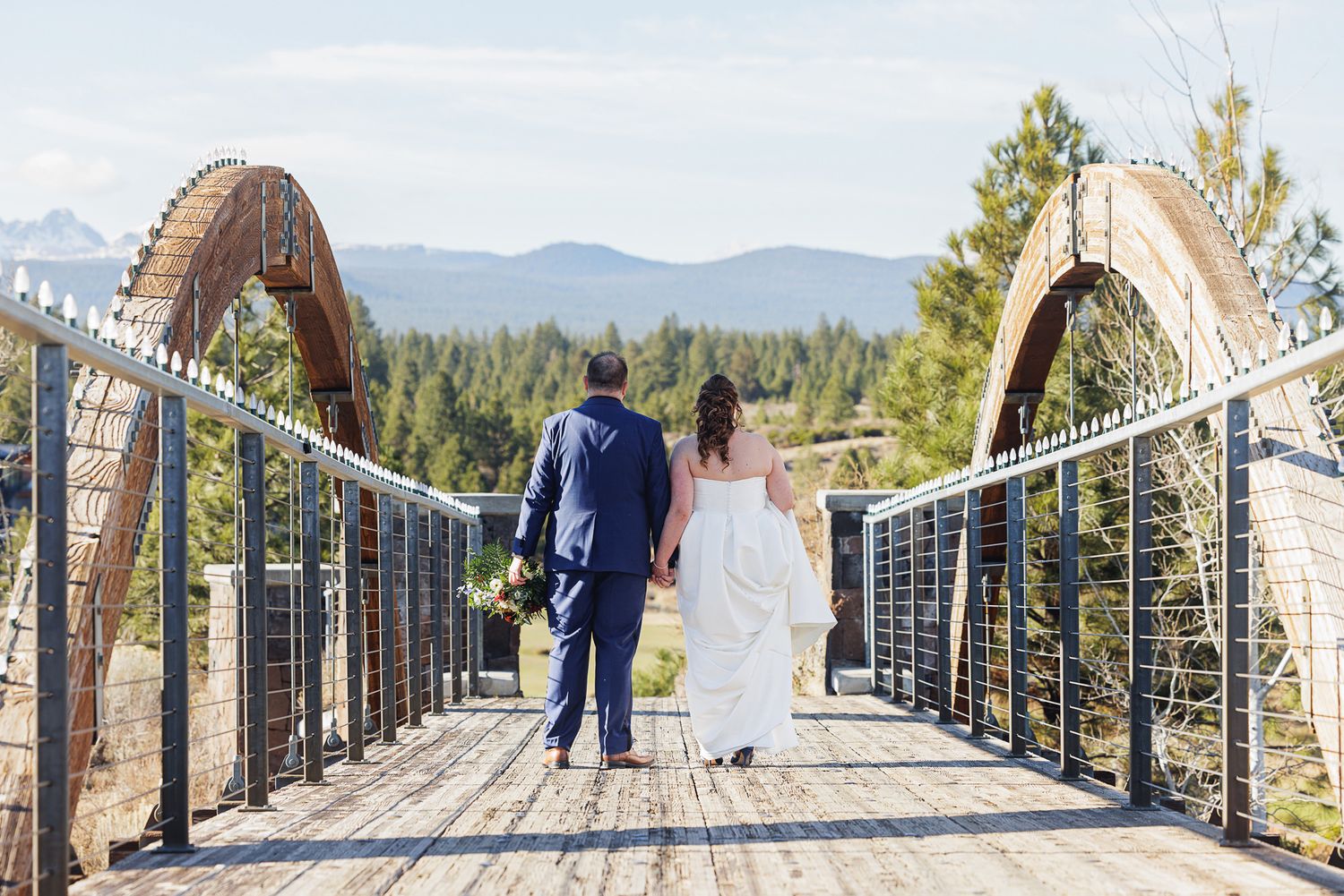 A couple walks across a scenic mountain footbridge surrounded by evergreen trees and distant mountain views.