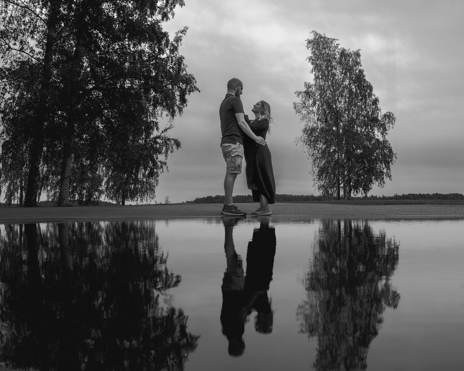 A couple stands silhouetted on a hill with their reflection mirrored in the calm lake water below, surrounded by trees.