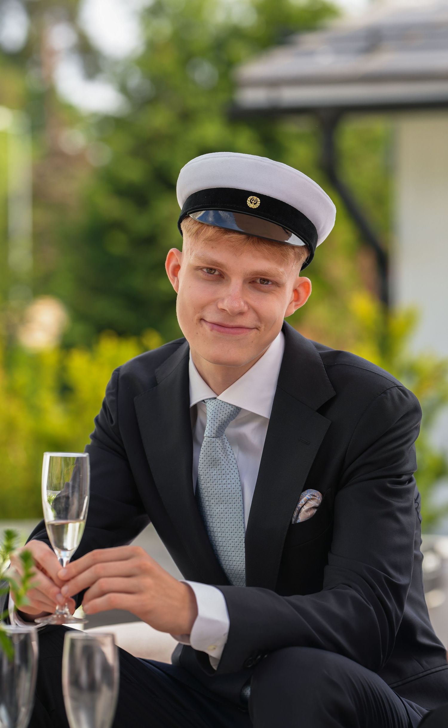 Person in formal attire and student cap holds champagne glass at outdoor celebration.