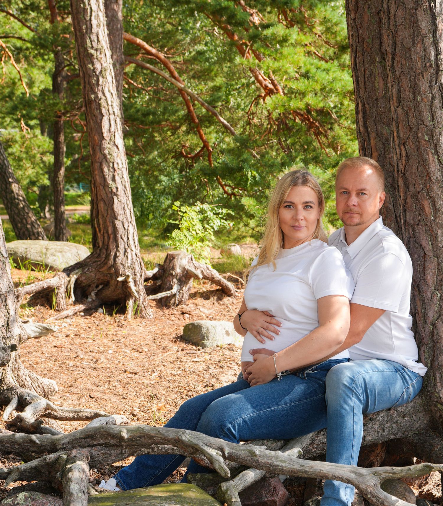Couple in white shirts sitting together on tree roots in a forest setting.