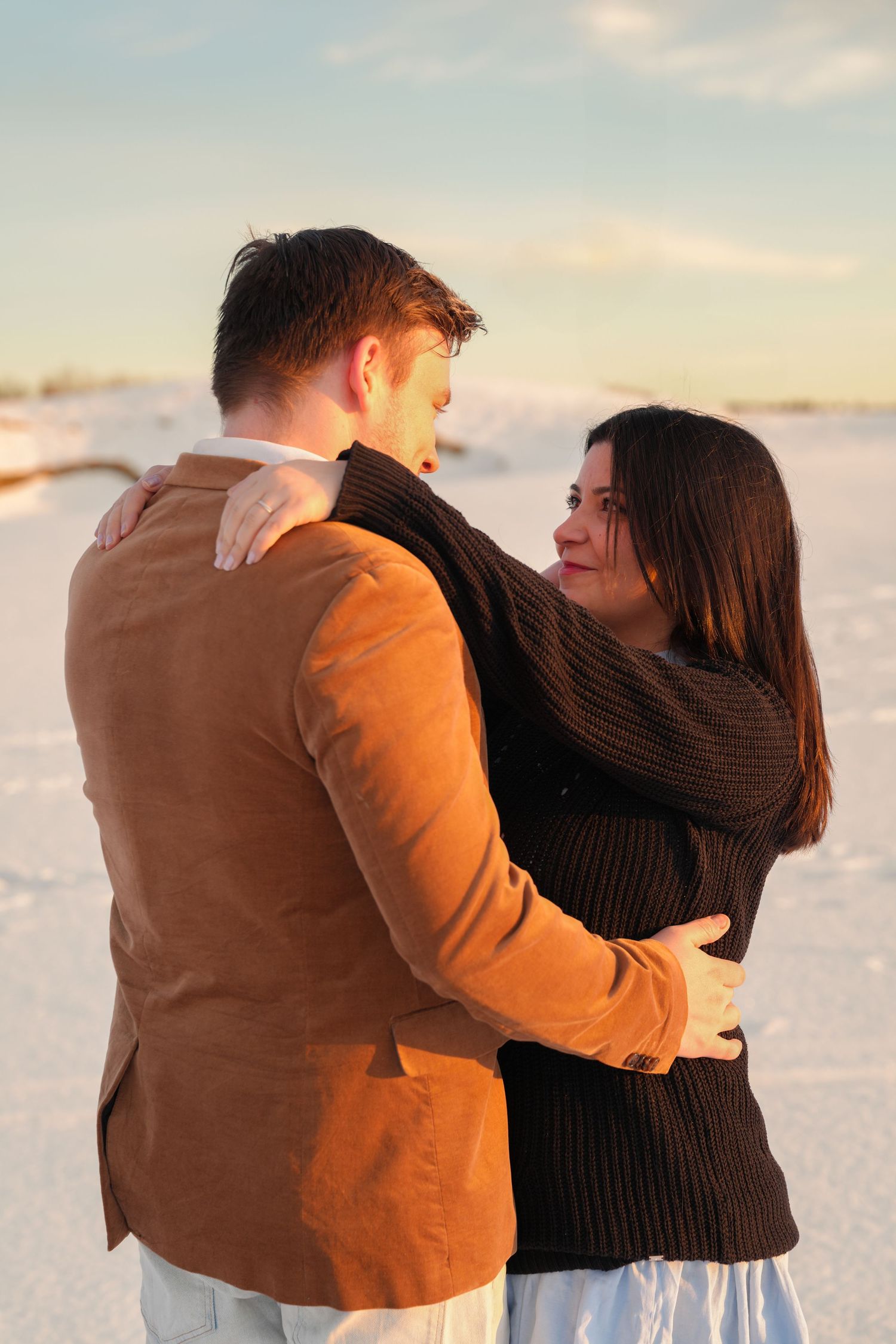 Couple in winter clothing embracing during golden hour in snowy setting.
