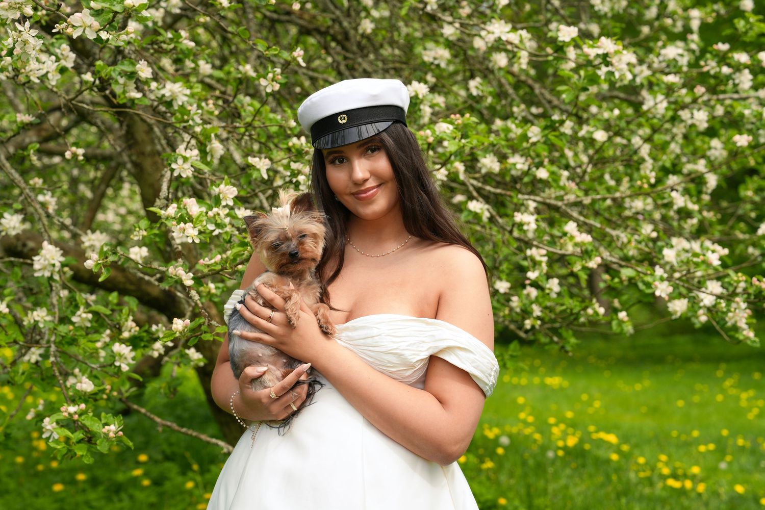 A woman in a white dress cuddles a small dog amidst blooming white flowers in a spring garden setting.