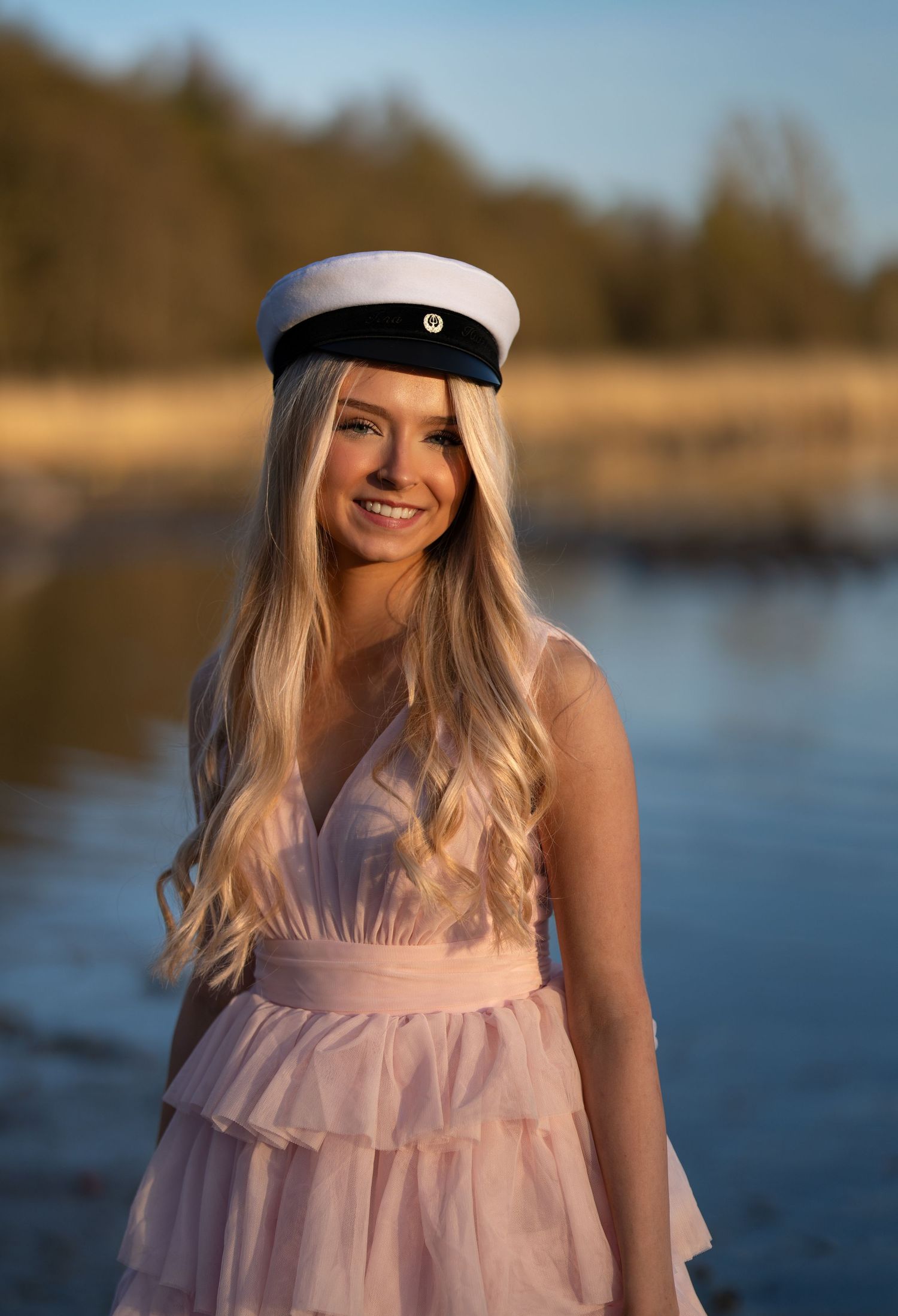 Smiling blonde woman in pink ruffled dress and white sailor cap posing outdoors near water at golden hour.