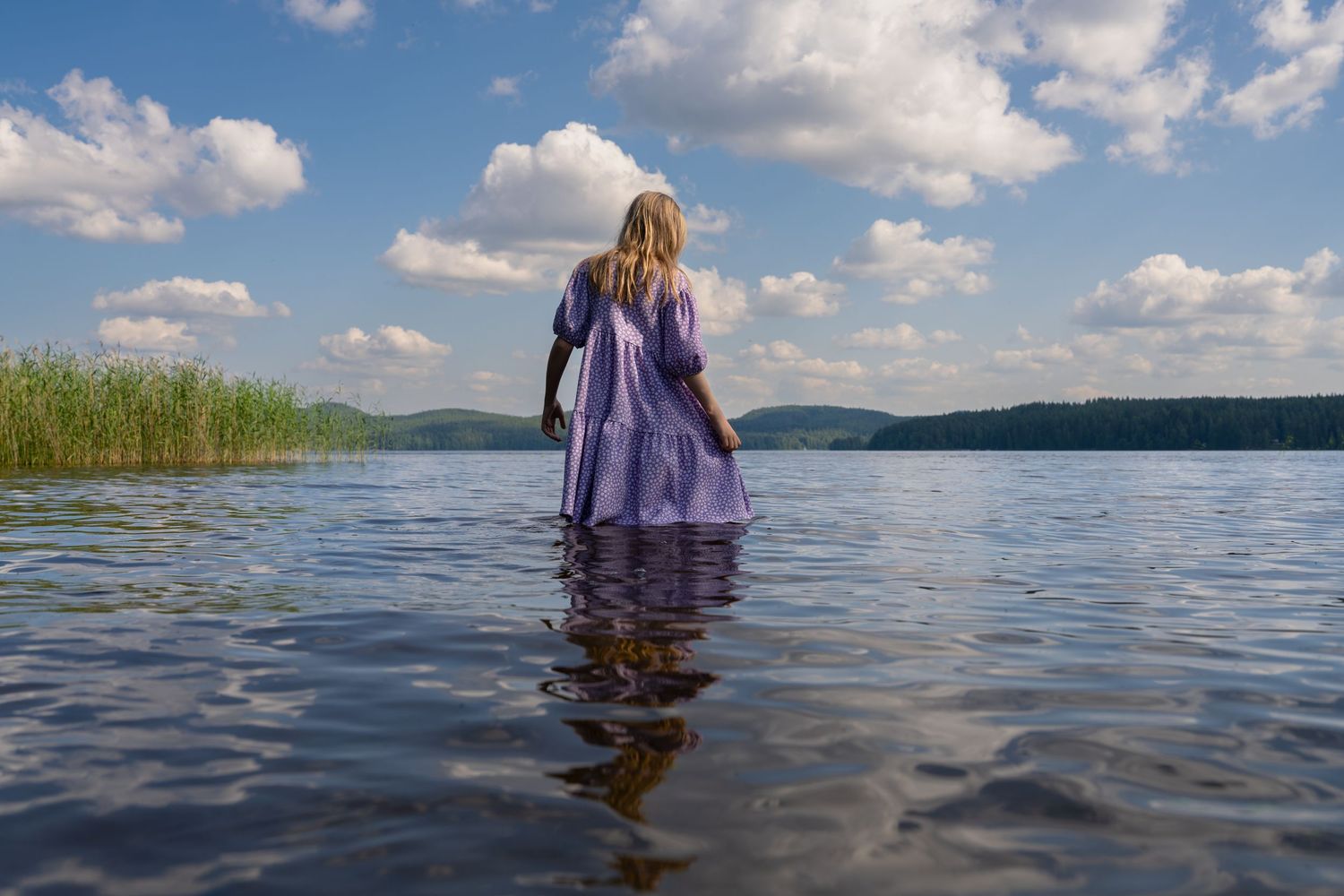 Figure in a purple dress wades into a serene lake surrounded by mountains and reeds on a sunny summer day.