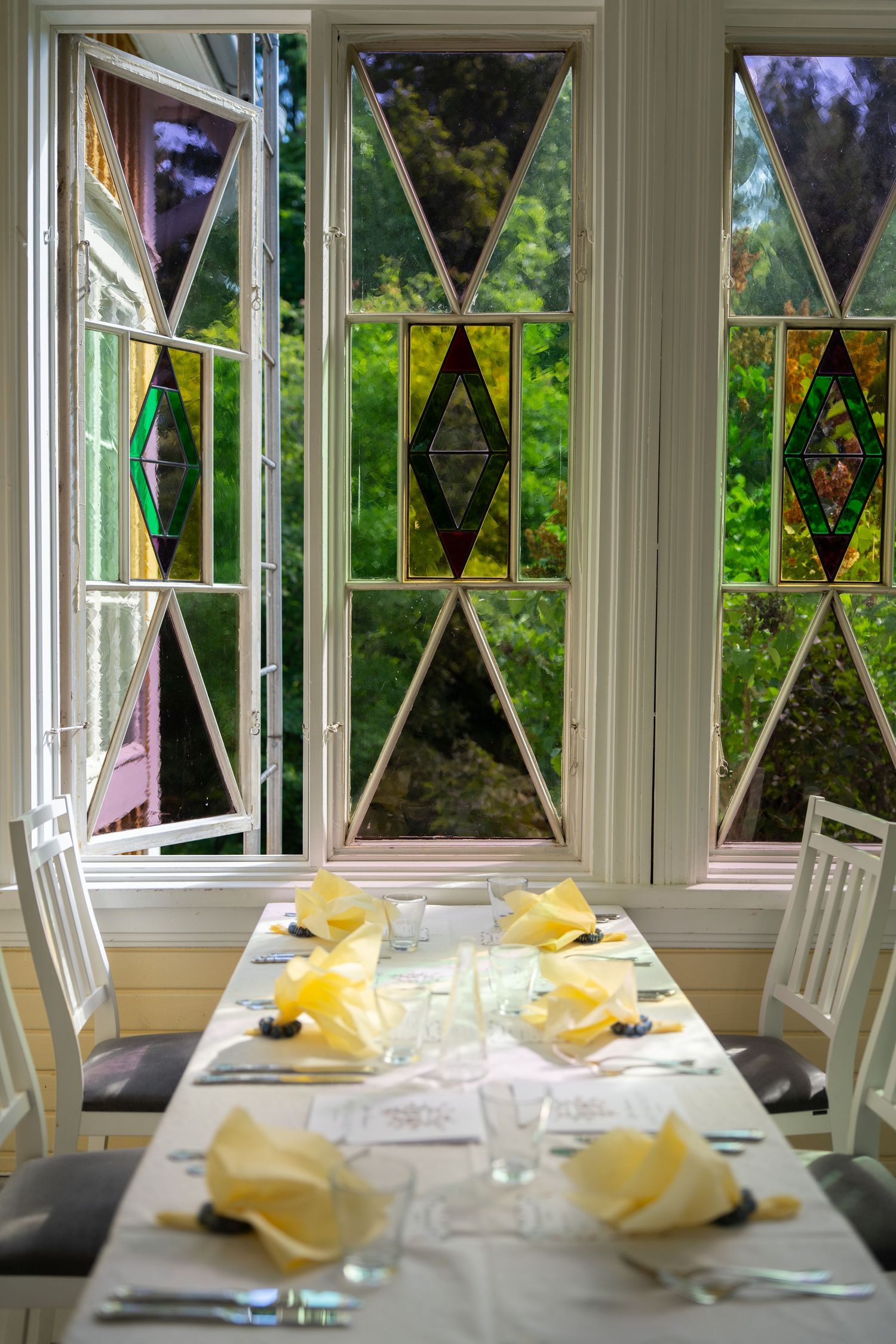 Elegant dining table set with yellow napkins near white-framed windows with colorful stained glass diamond panes overlooking lush greenery.