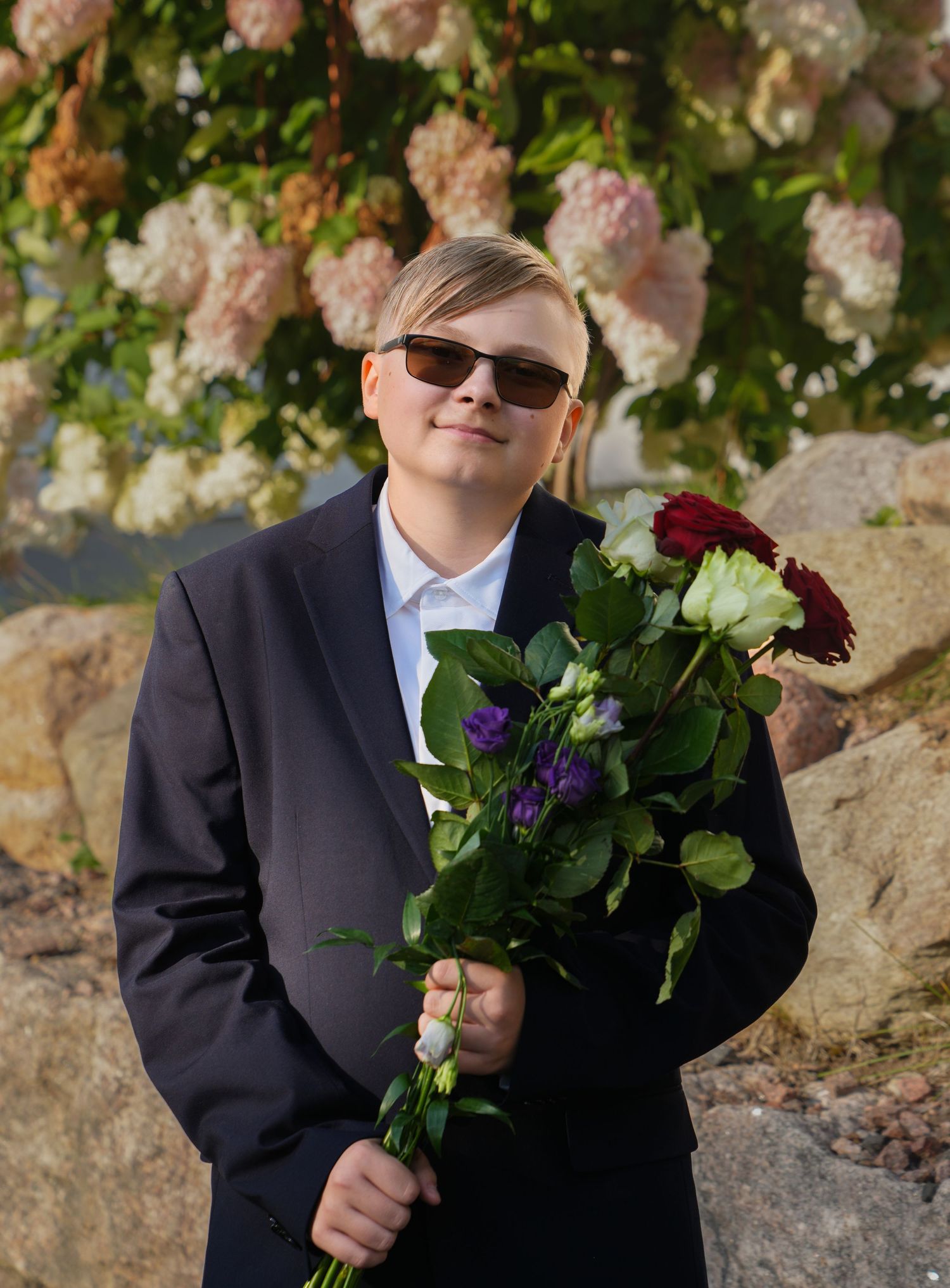 Person in dark suit holding colorful bouquet of roses against flowering bush backdrop.