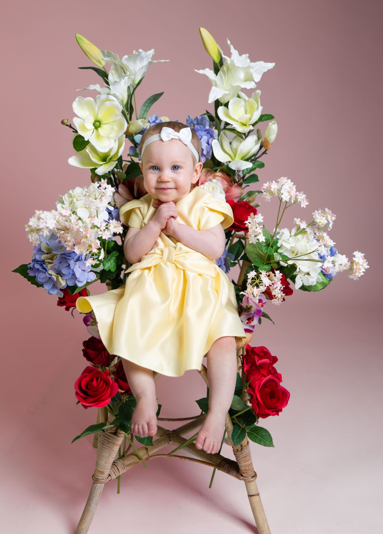 Baby girl in yellow dress sits on floral chair surrounded by colorful blooms on pink background.