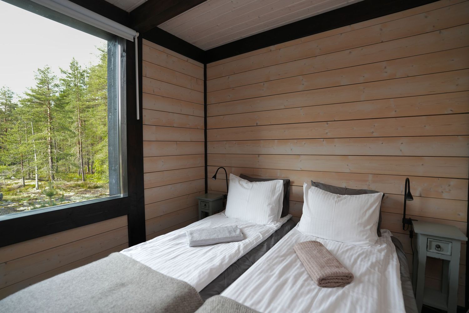 Cozy bedroom with twin beds and wooden walls featuring a large window overlooking a pine forest.
