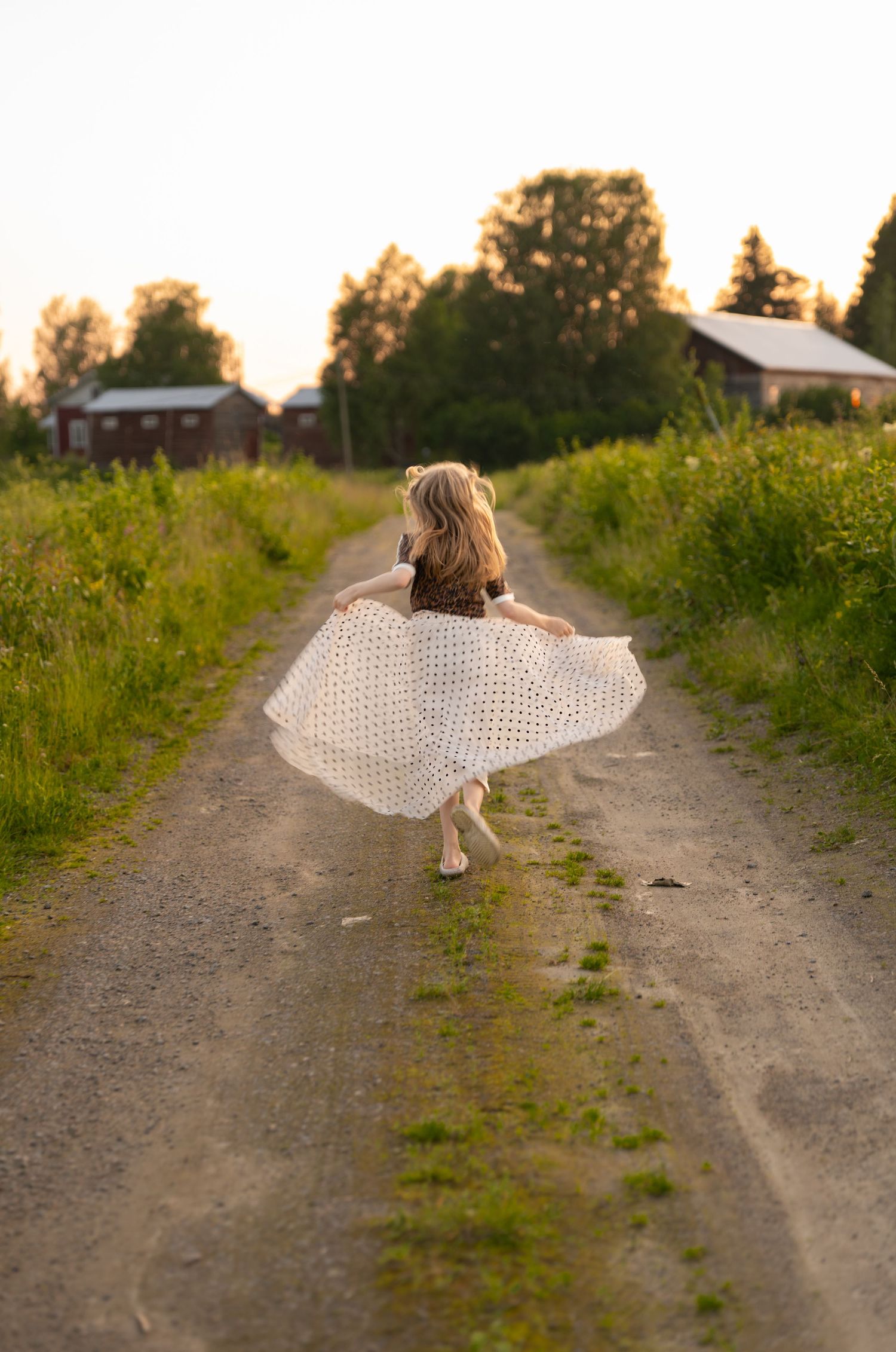 A figure in a flowing white dress twirls on a rural dirt path at sunset with farmhouses in the background.