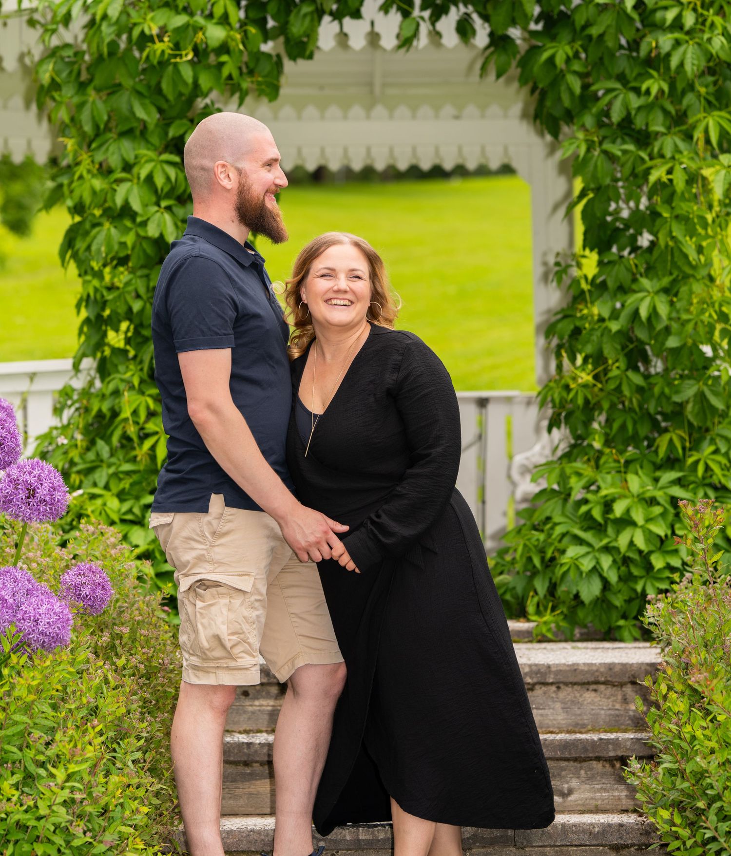 A couple shares a joyful moment laughing together on garden steps near a white arbor with purple flowers and greenery.
