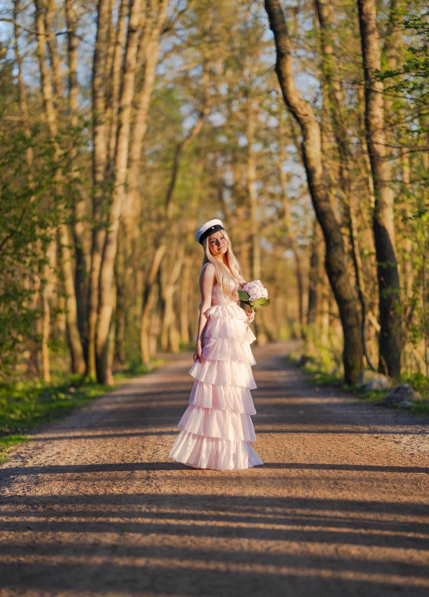 Bride in pink tiered gown holding bouquet stands on tree-lined path in golden sunlight.