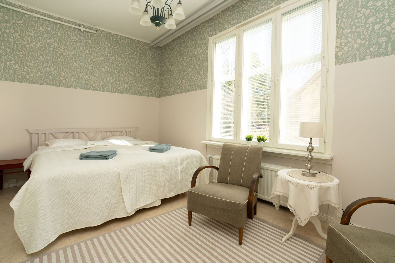 Elegant bedroom with floral wallpaper, white bedding, striped rug, and natural light from large windows.