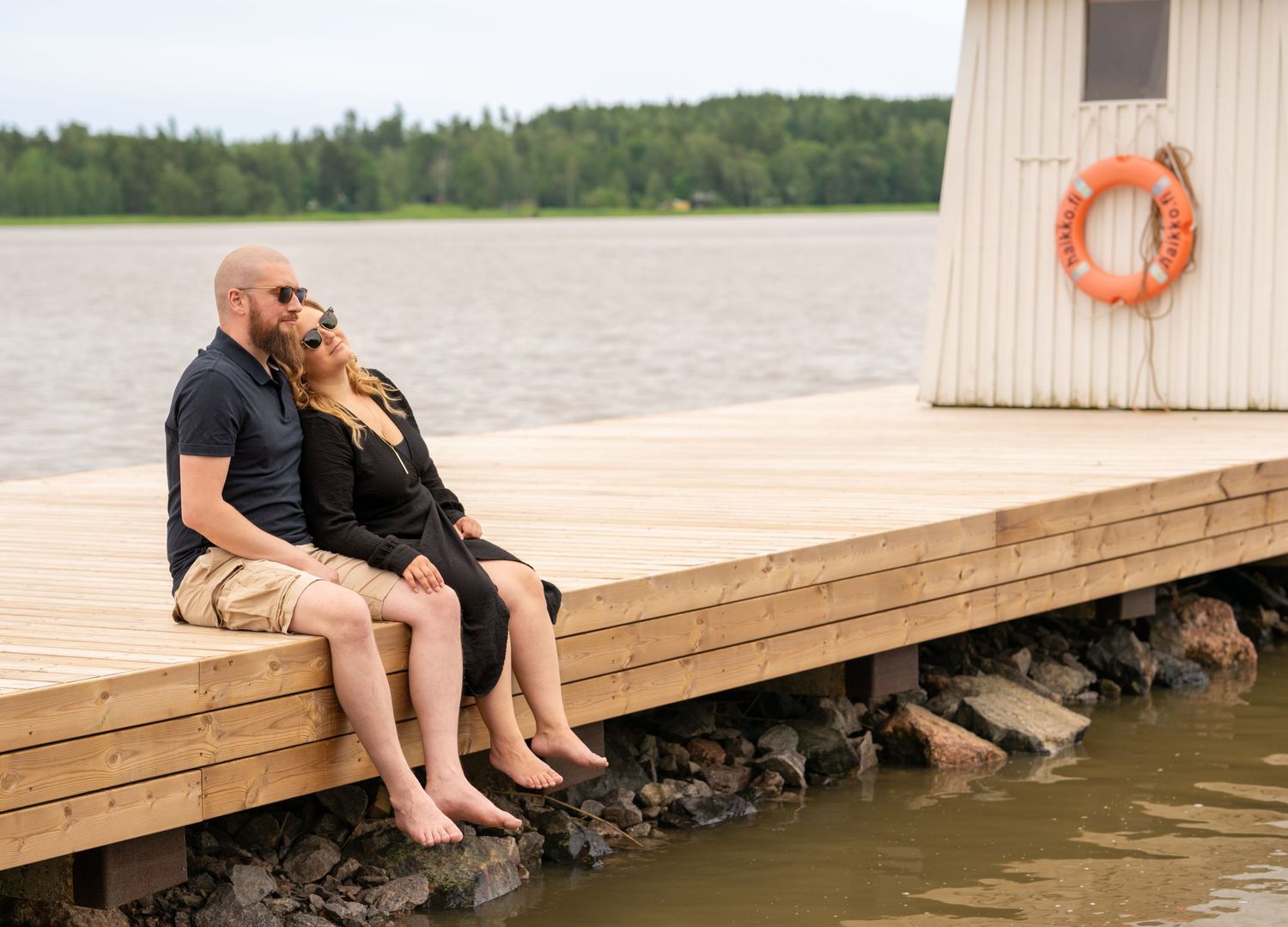 A couple sits together on a wooden dock by the water with a life preserver on the wall behind them.