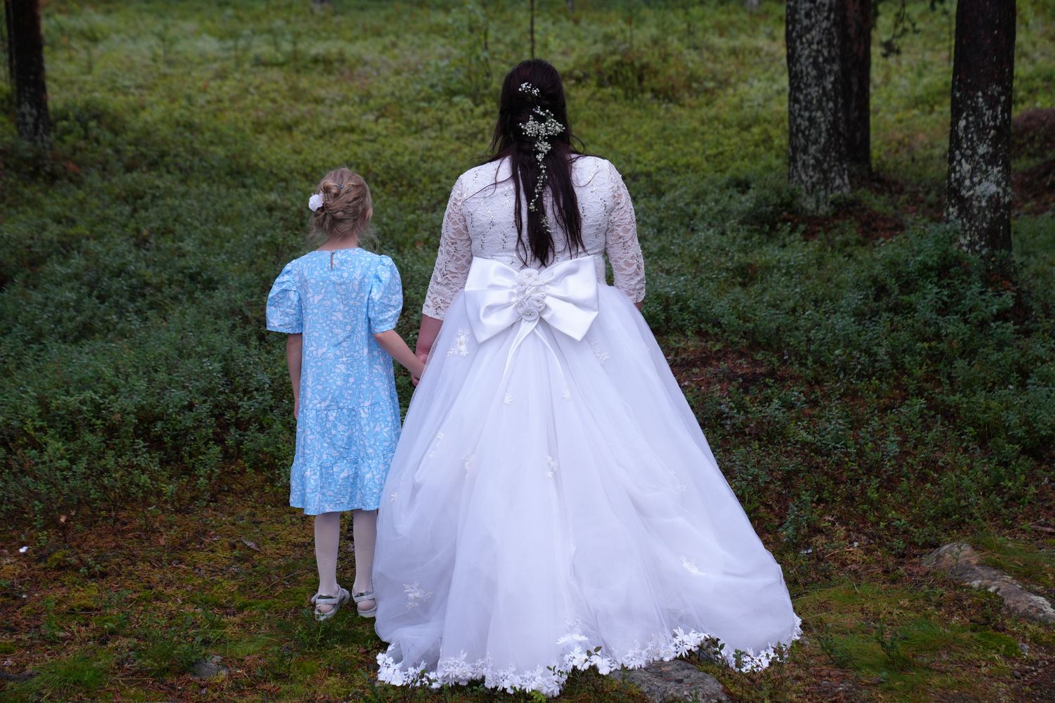 Back view of a bride in a white gown with bow detail standing with small attendant in blue dress in forest setting.