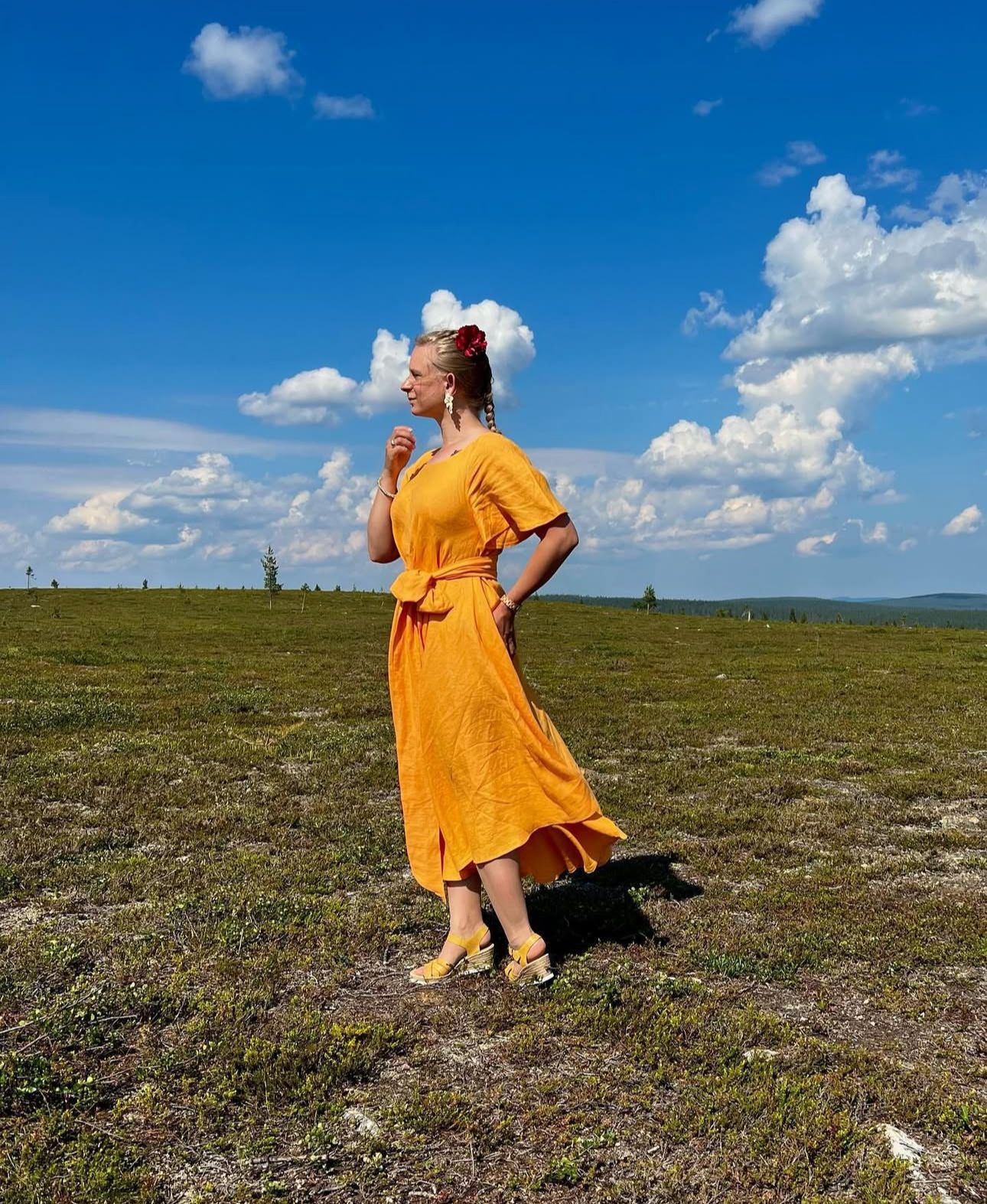 Woman in flowing yellow dress standing in a grassy field under puffy white clouds.